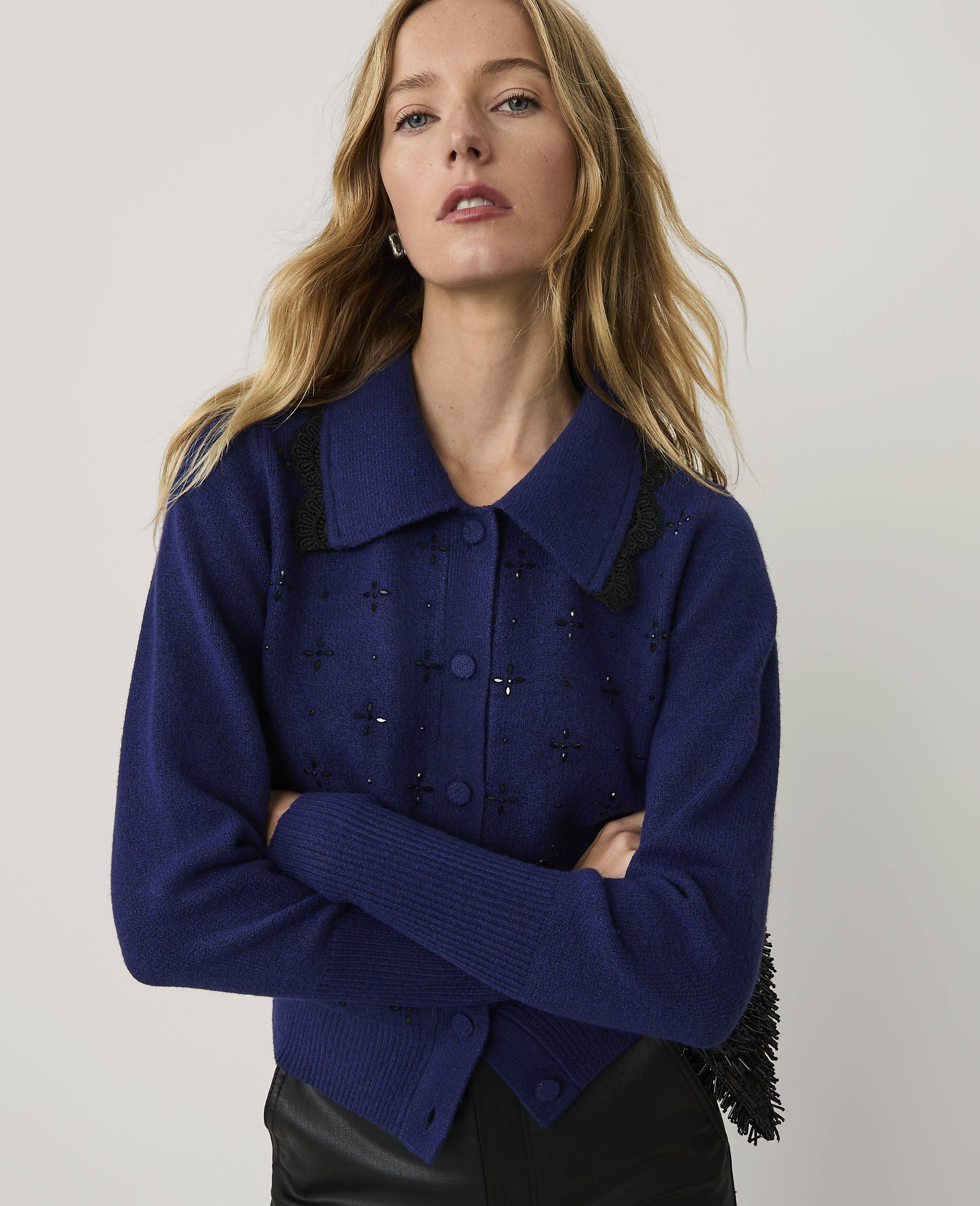 A blonde woman wearing a blue embellished cardigan and black leather pants stands against a plain white background.