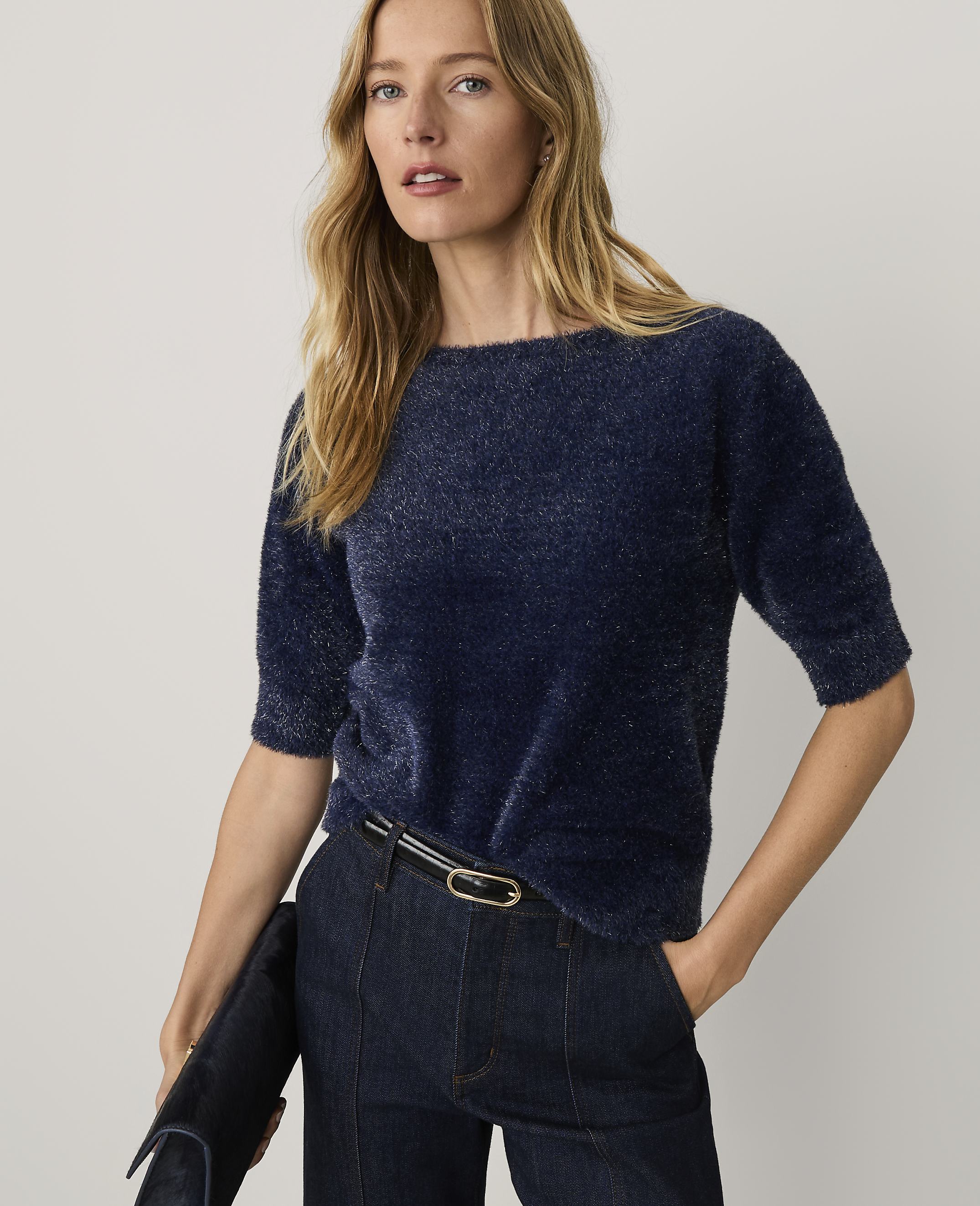A woman wearing a navy blue fuzzy sweater and dark jeans stands against a plain, light-colored background.