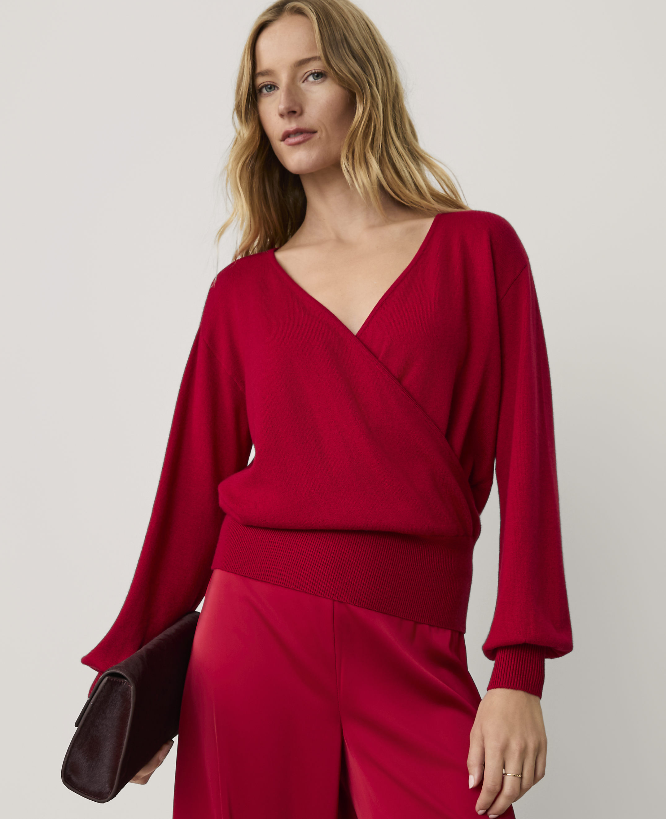 A woman wearing a red wrap-style top and matching red trousers holds a dark clutch against a plain, light-colored background.