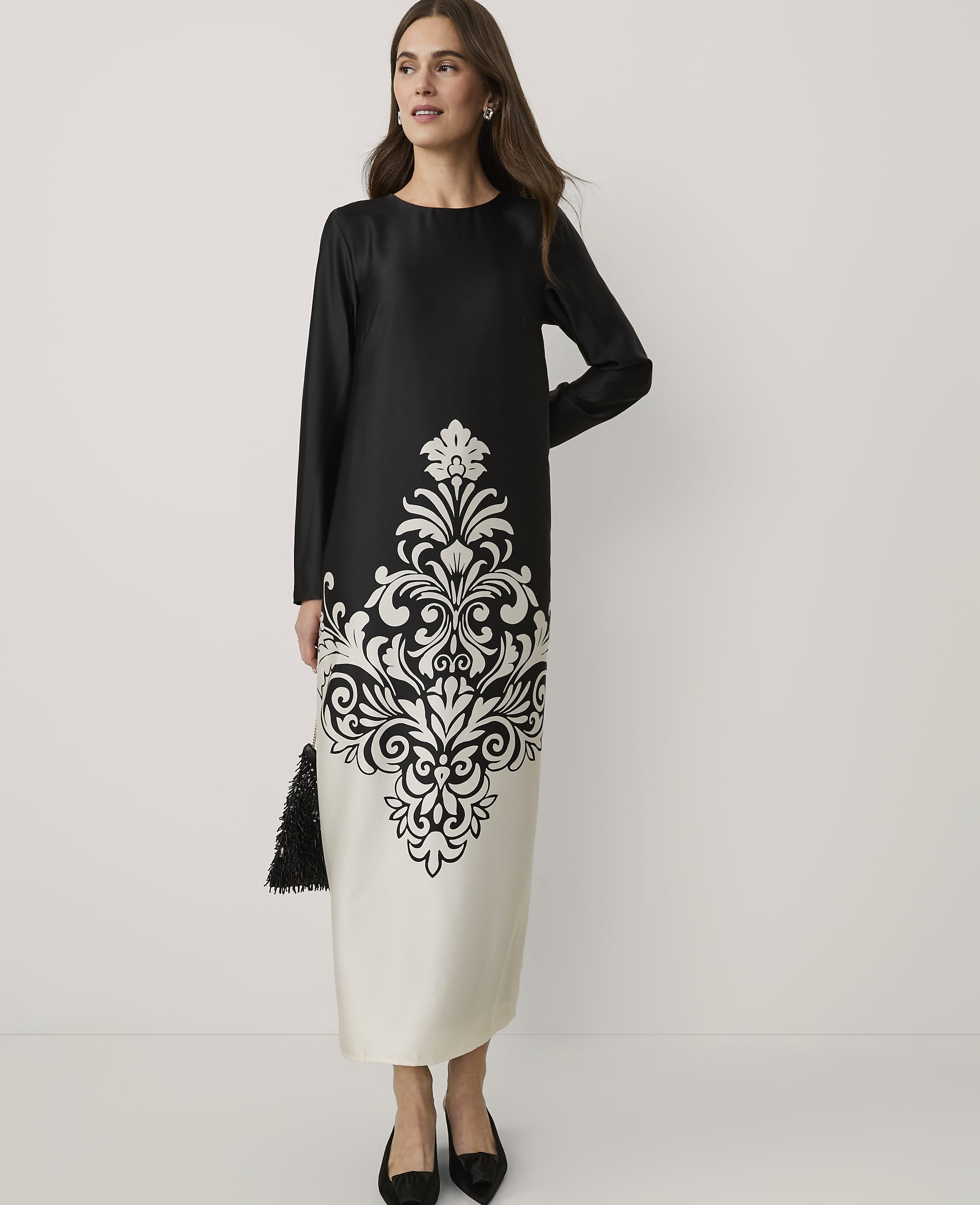 A woman wearing a black and white patterned midi dress and black heels stands against a plain white background.
