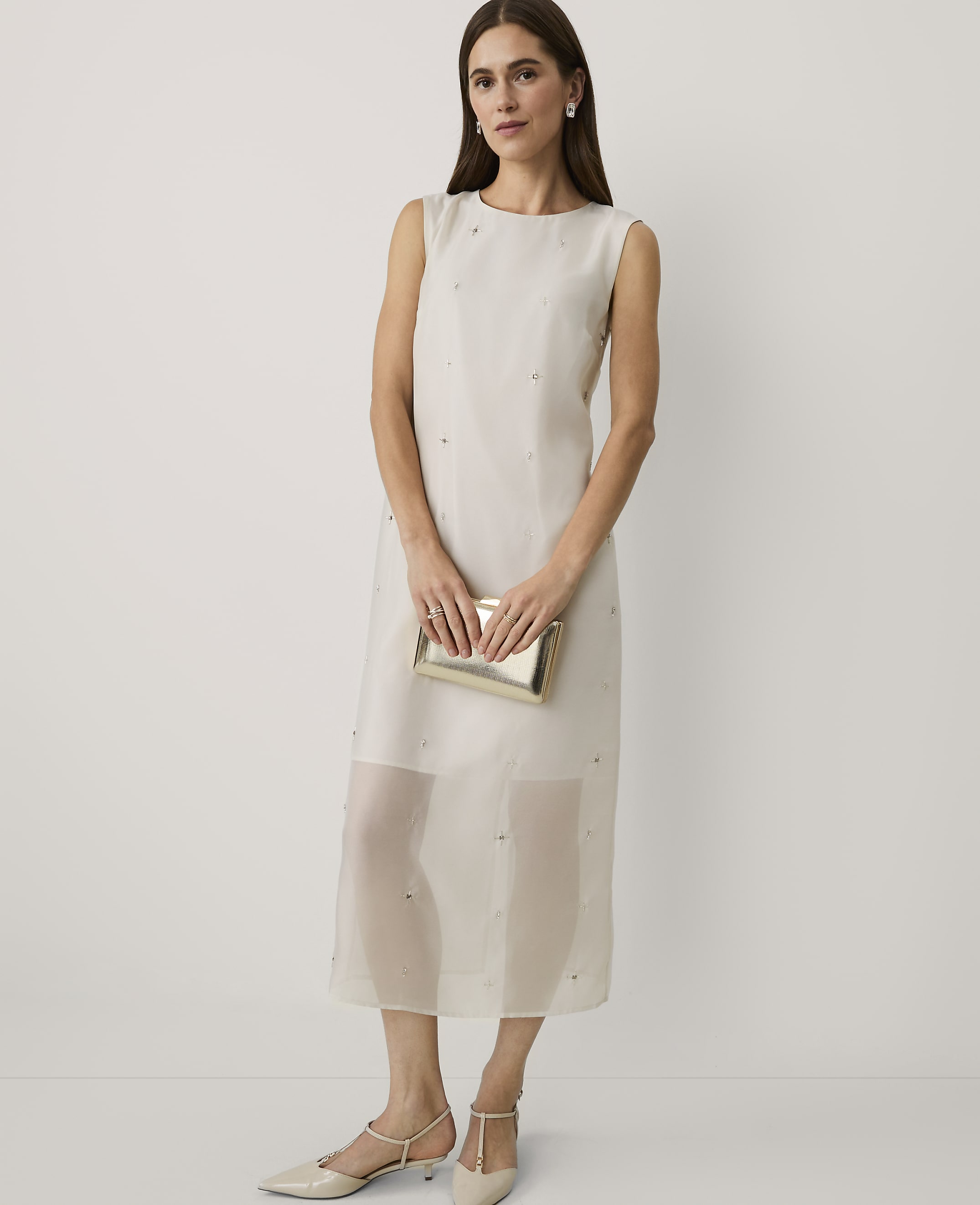 A woman wearing a sleeveless, embellished cream dress and heels holds a gold clutch against a plain white background.