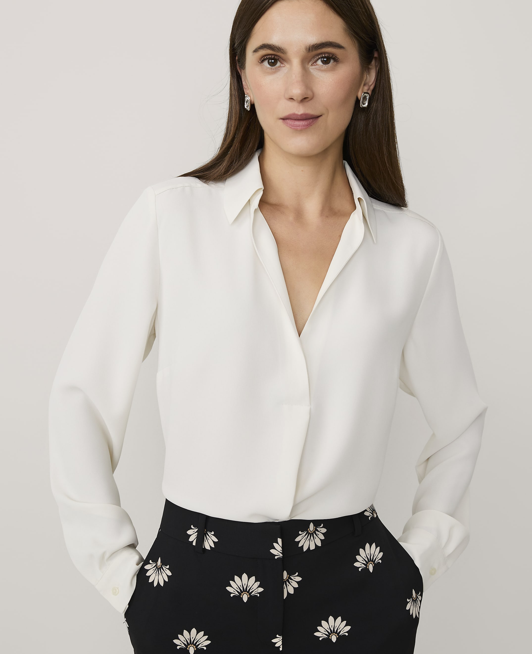 A woman wearing a white long-sleeved blouse and black floral trousers stands against a plain light gray background.