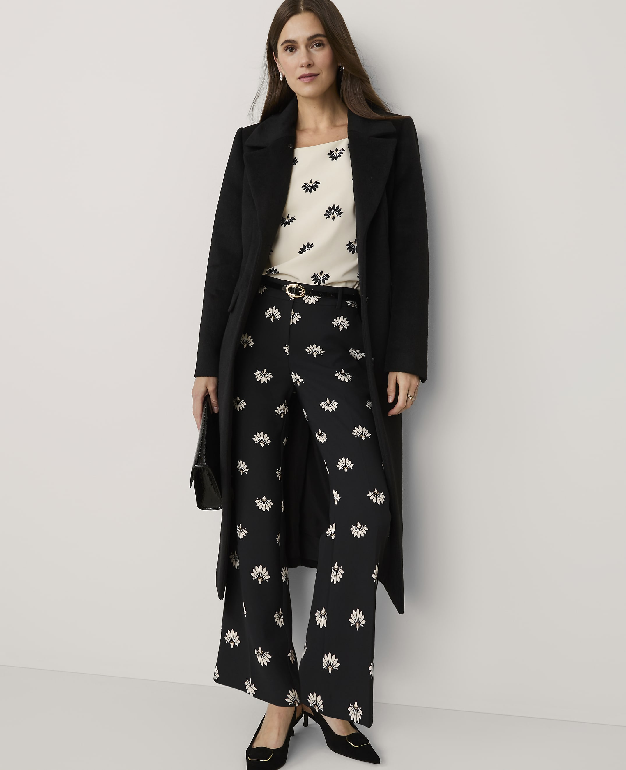 A woman wearing a black coat, a floral top, and matching floral trousers stands against a plain white background.