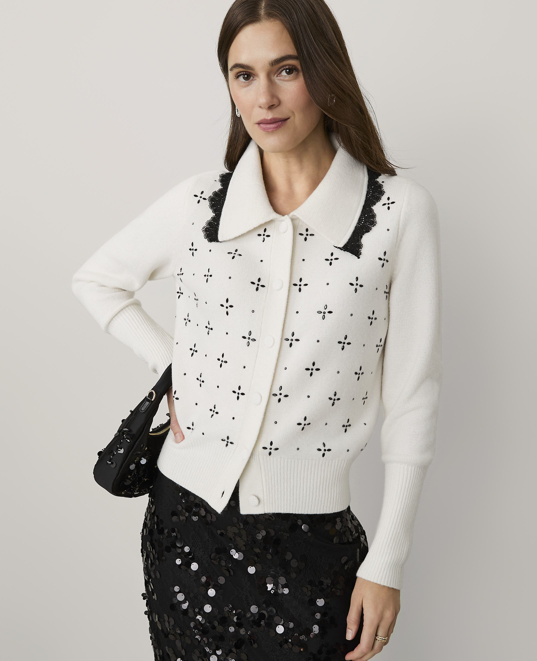 A woman wearing a white embellished cardigan and a black sequined skirt stands against a plain light gray background.