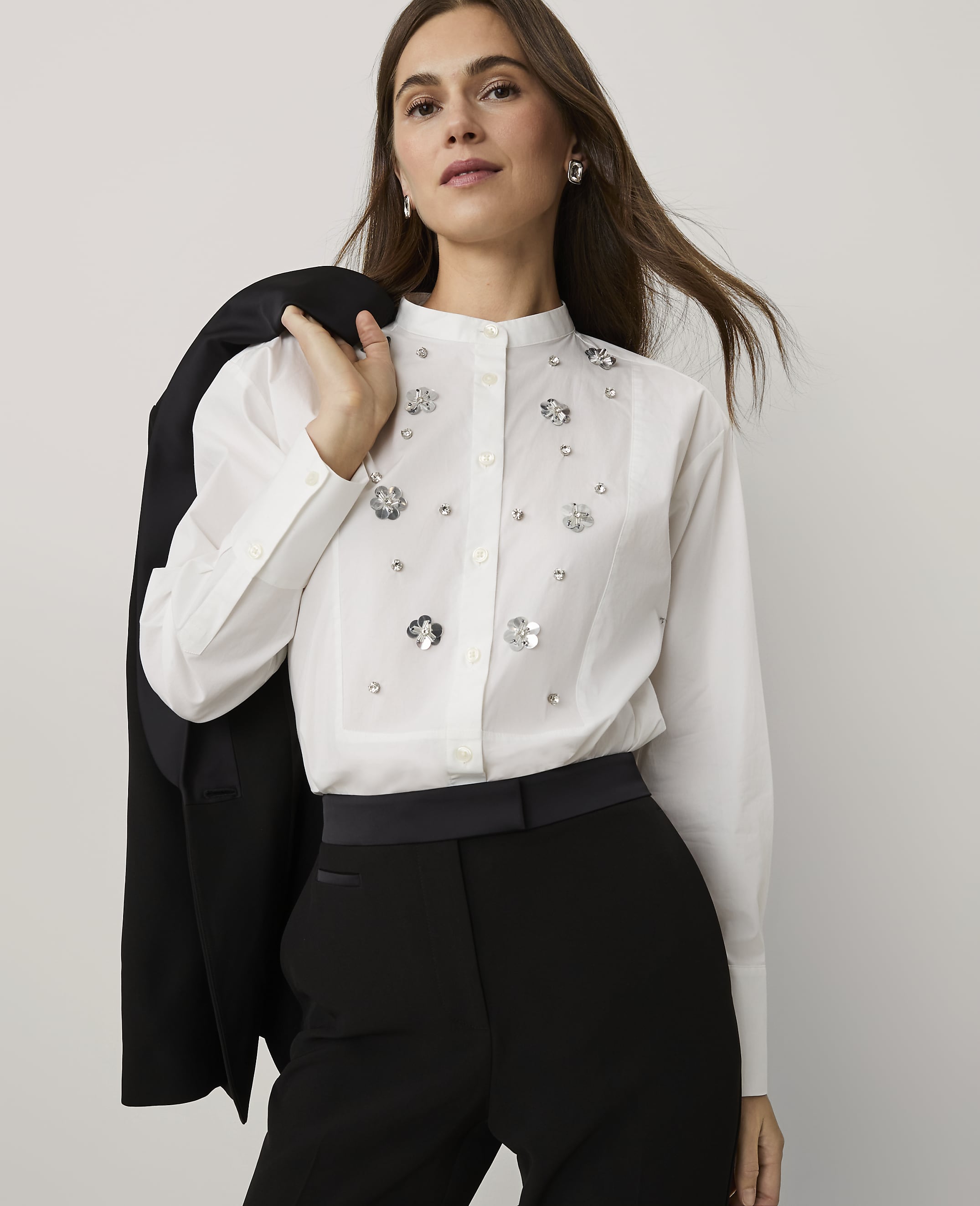 A woman wearing a white embellished shirt and black trousers stands against a plain light grey background, holding a black blazer over her shoulder.