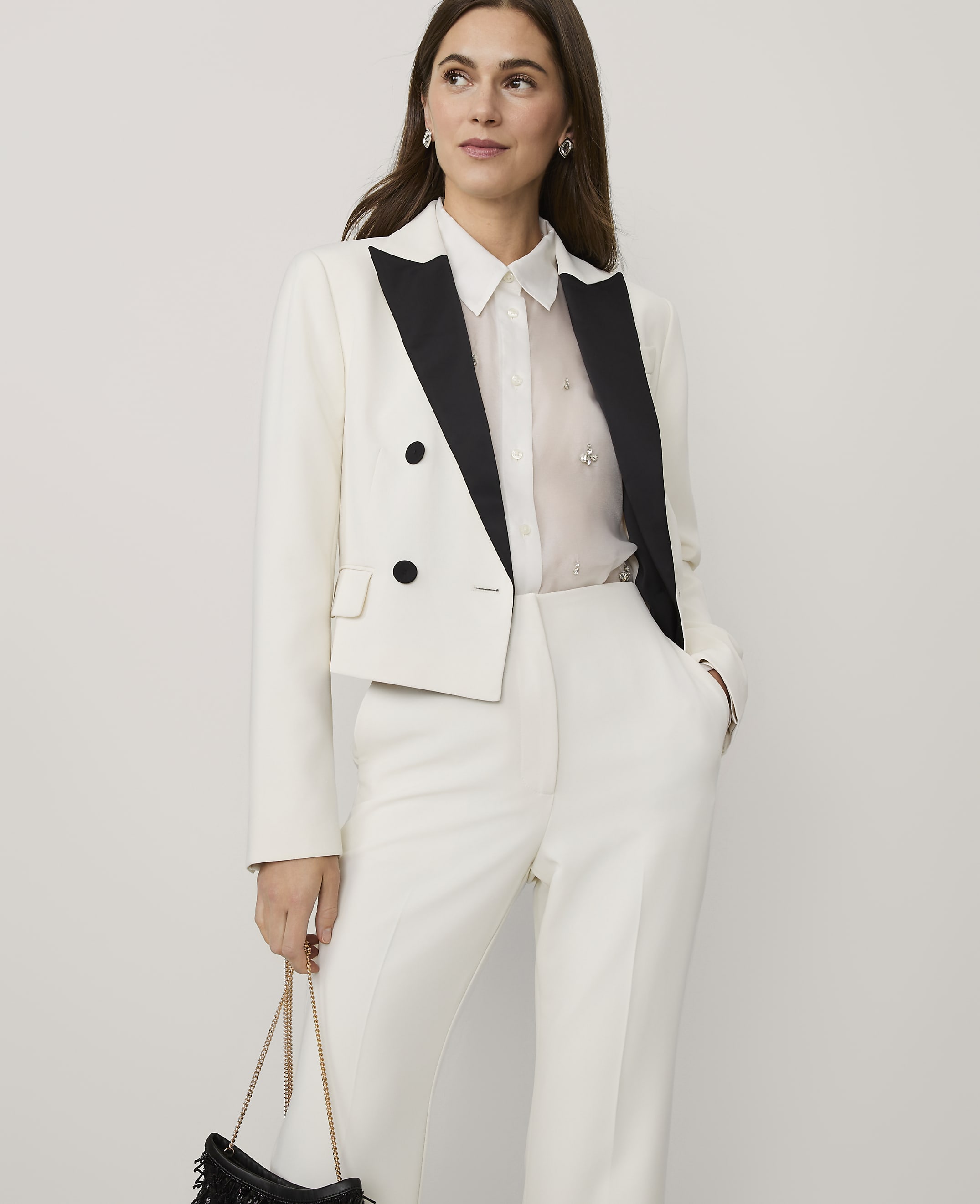A woman wearing a white cropped blazer and matching trousers stands against a plain, light-colored background.