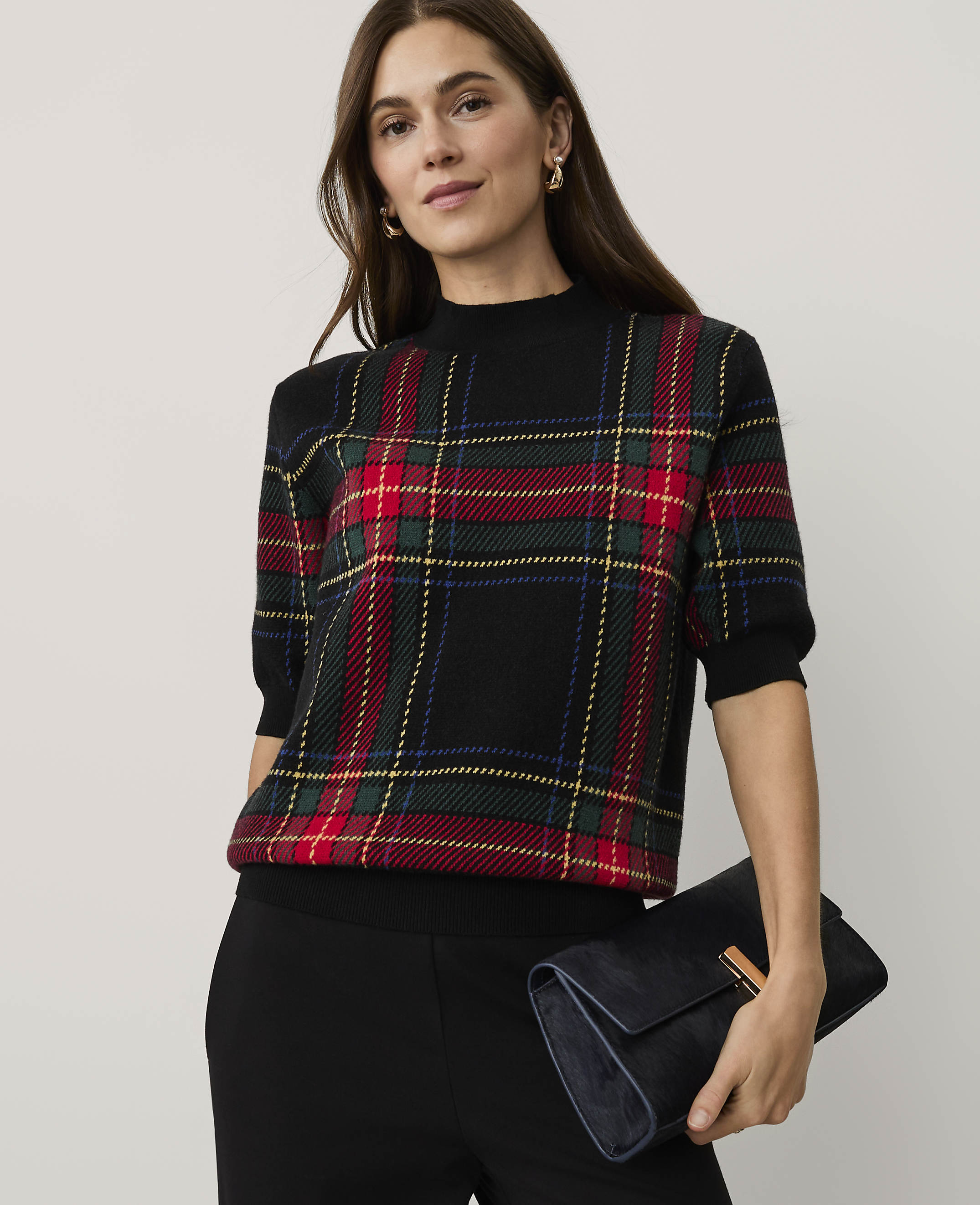 A woman wearing a red and green plaid sweater and black pants holds a dark blue clutch against a plain light grey background.