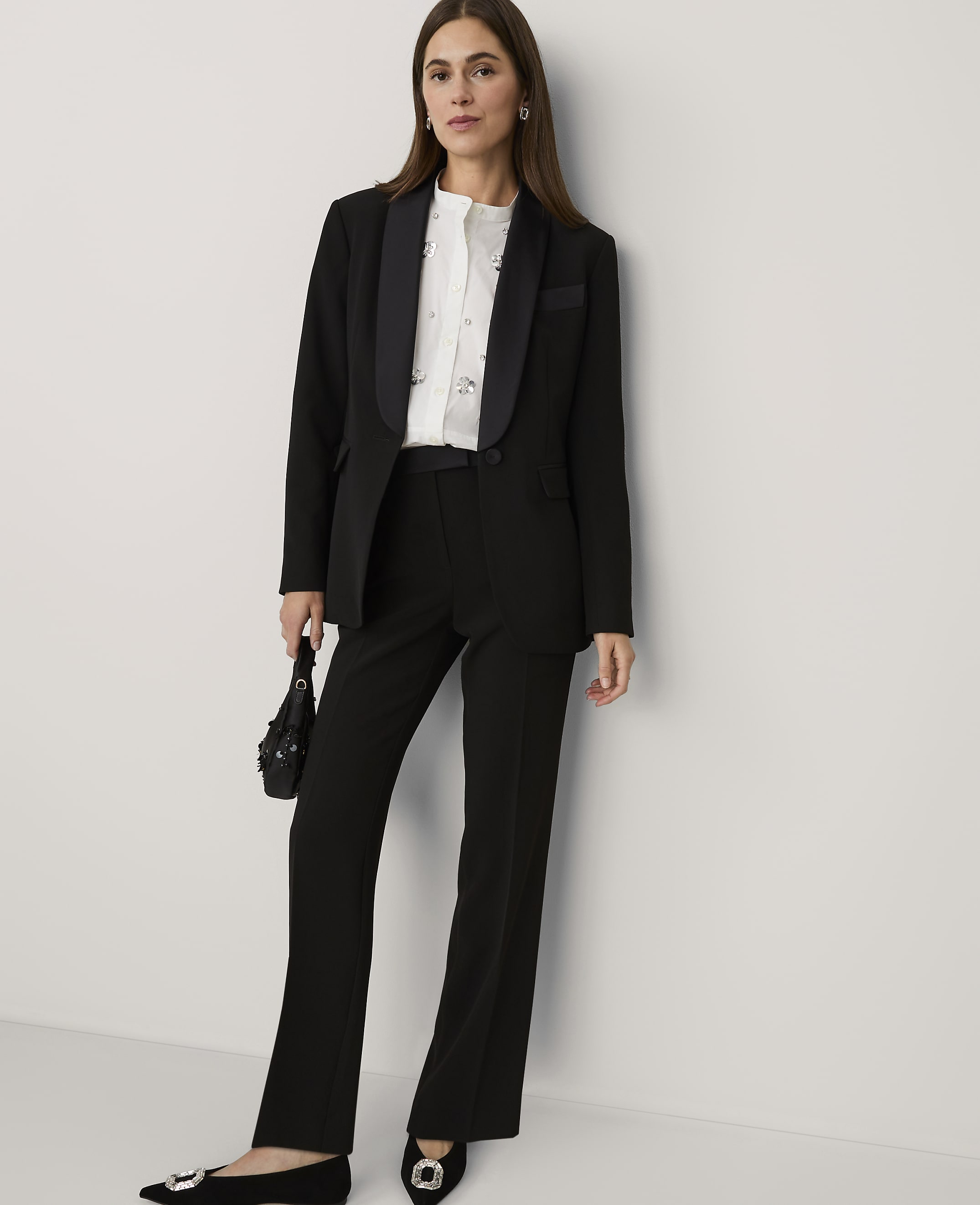 A woman wearing a black suit and white blouse stands against a plain white wall.