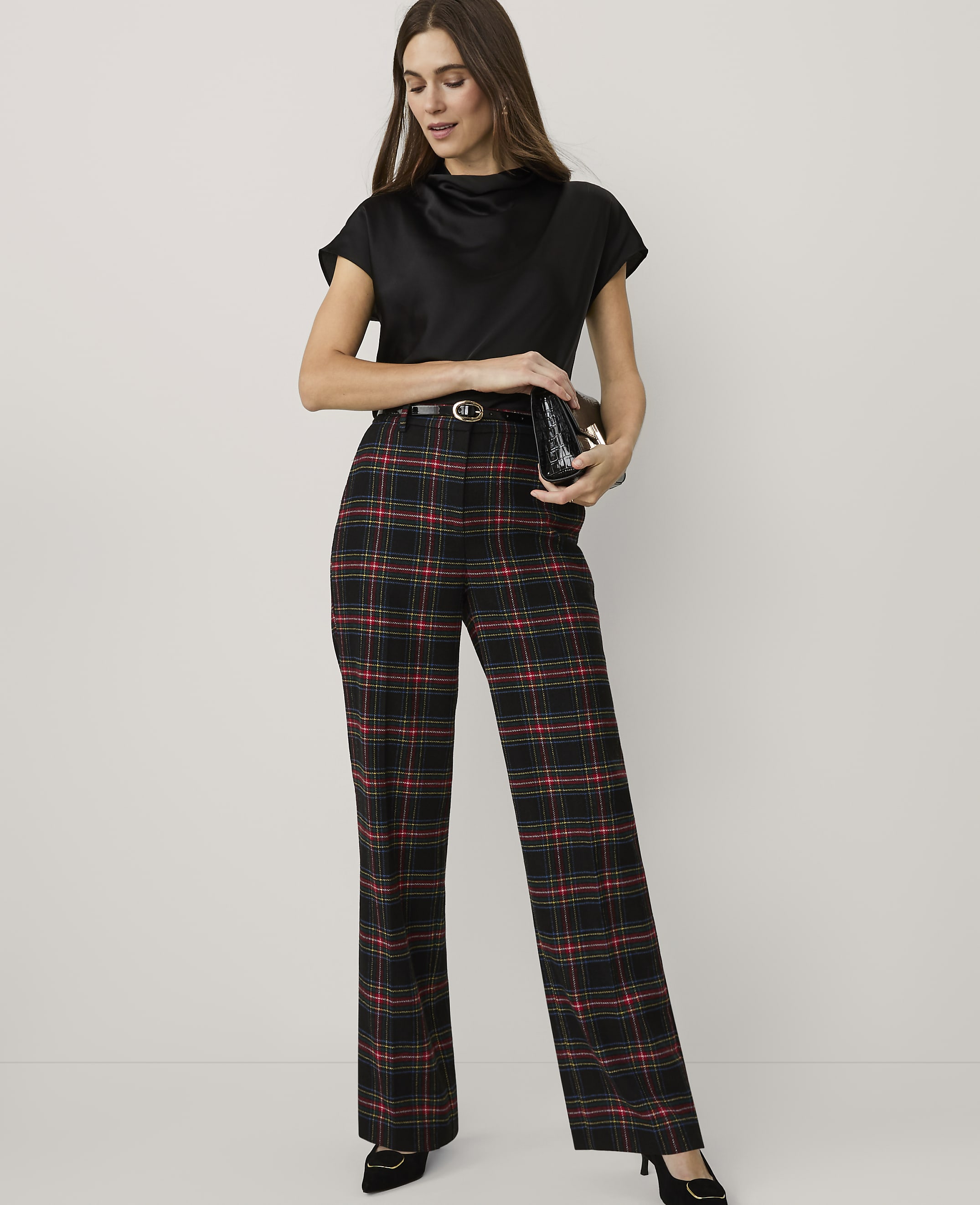 A woman wearing a black top and red and black plaid trousers stands against a plain white background.