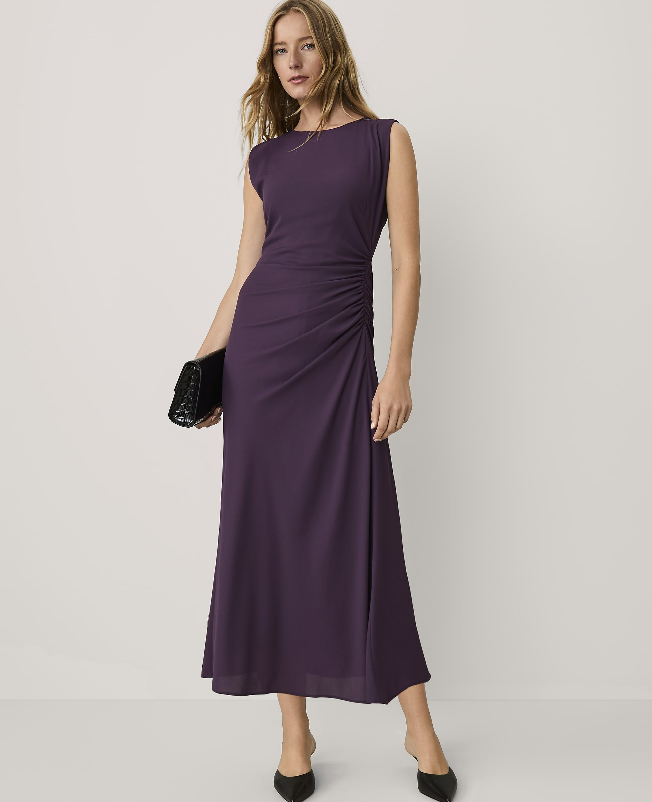 A woman wearing a sleeveless purple maxi dress and black heels stands against a plain white background.