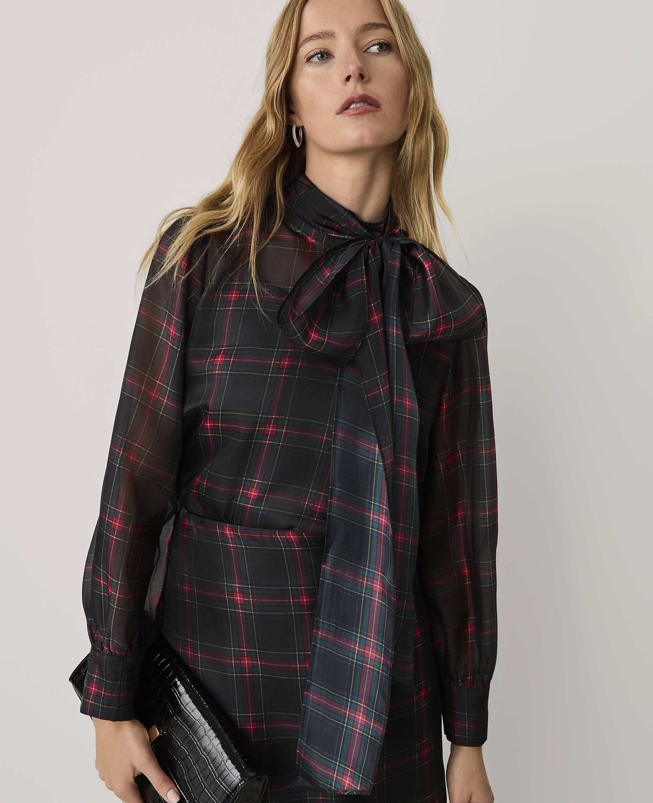 A woman wearing a sheer black and red plaid blouse with a pussy-bow neckline stands against a plain, light-colored background.