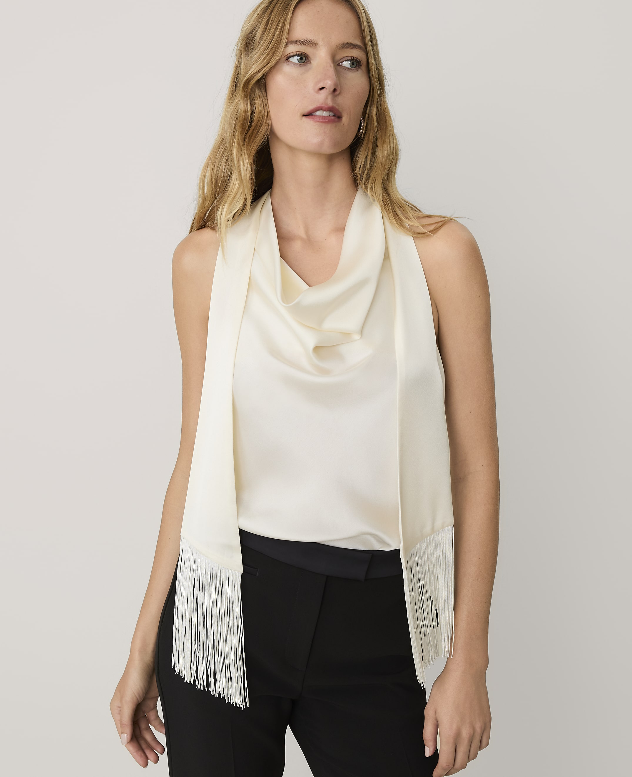 A woman wearing a cream-colored cowl-neck top and a matching fringed scarf stands against a plain light gray background.