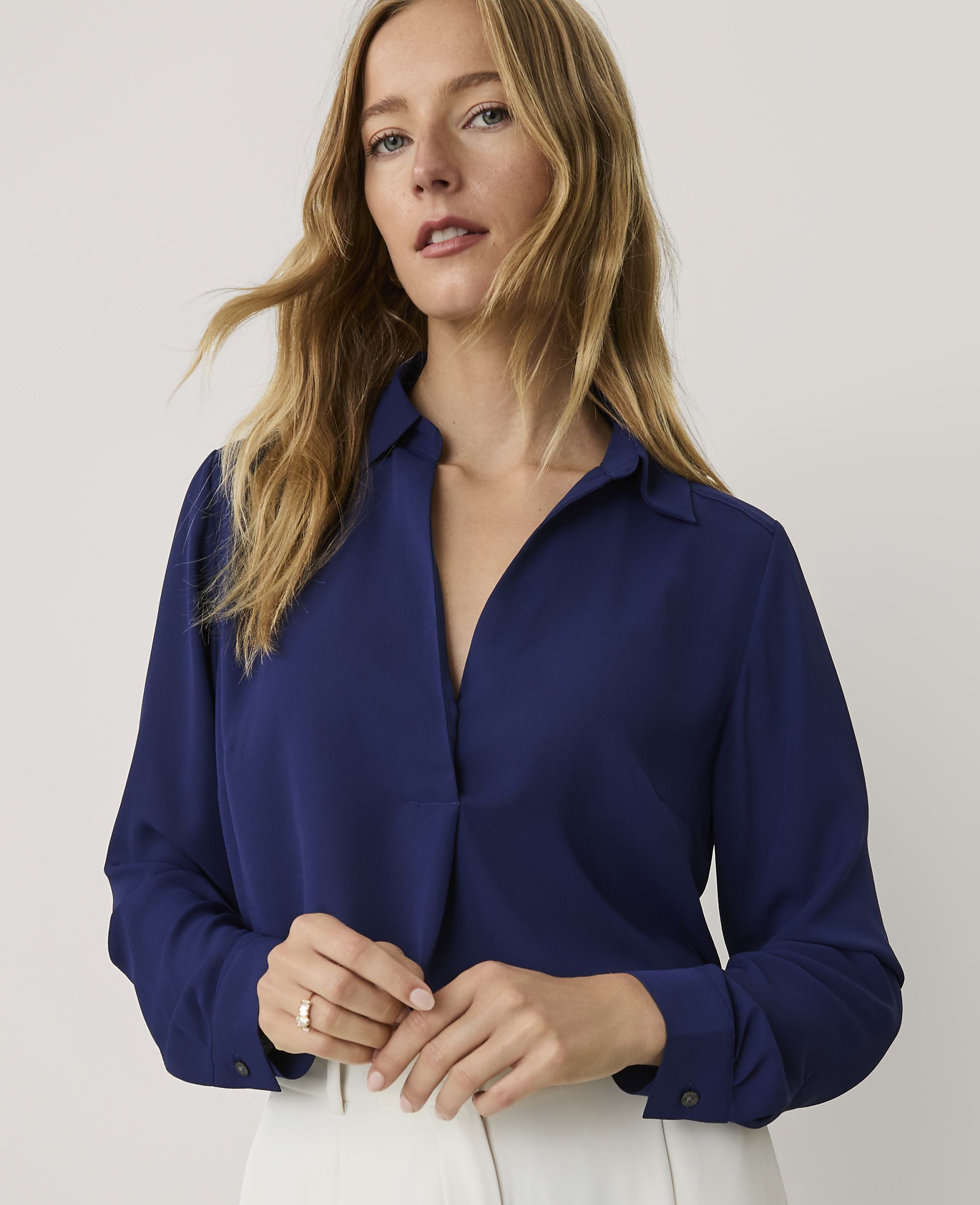 A blonde woman wearing a royal blue button-down shirt and white trousers stands against a plain, light-colored background.