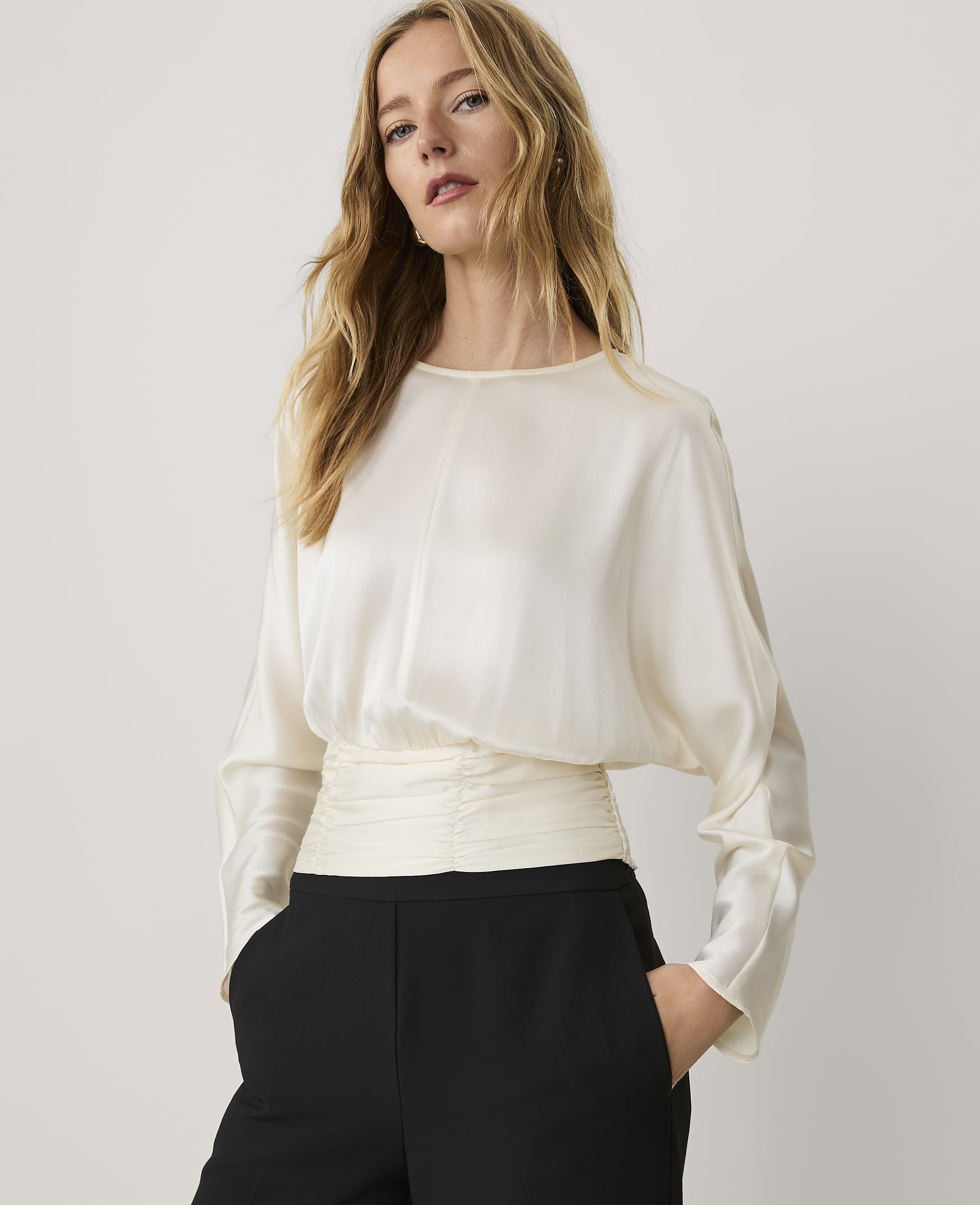 A woman wearing a cream-colored satin blouse and black trousers stands against a plain, light-colored background.