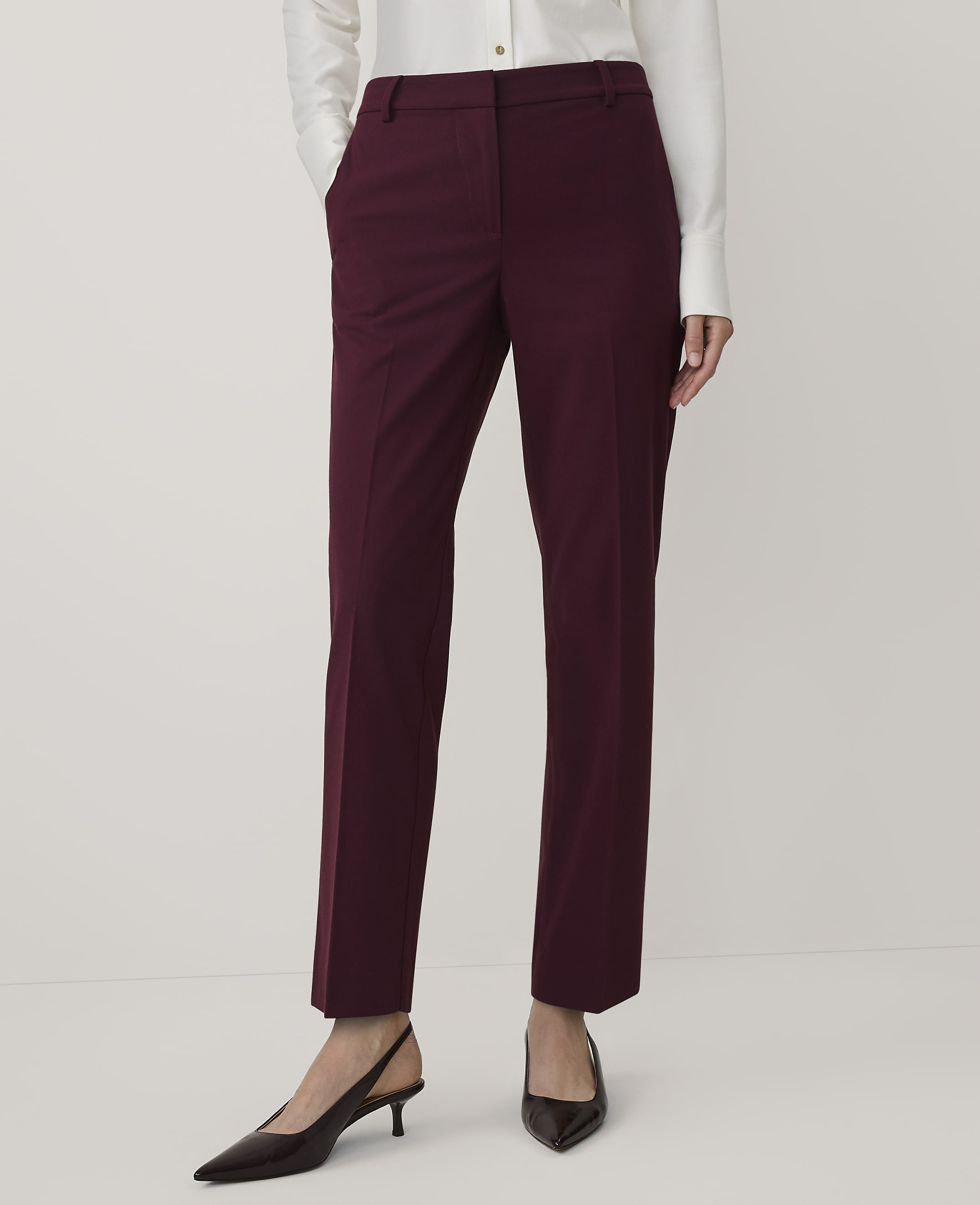 A person wearing a white shirt, burgundy trousers, and dark pointed-toe heels stands against a plain white background.