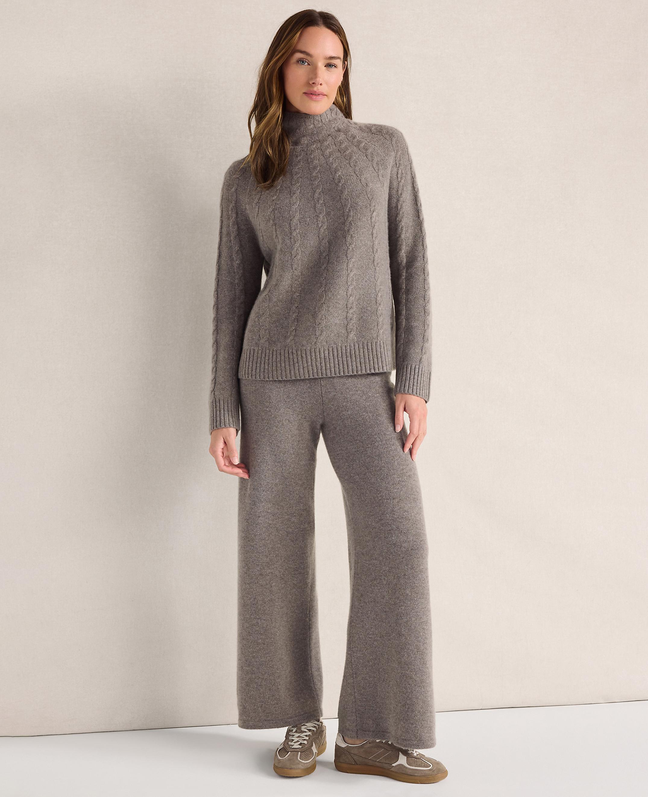 A woman wearing a grey cable-knit sweater and matching wide-leg trousers stands against a plain, neutral-colored background.