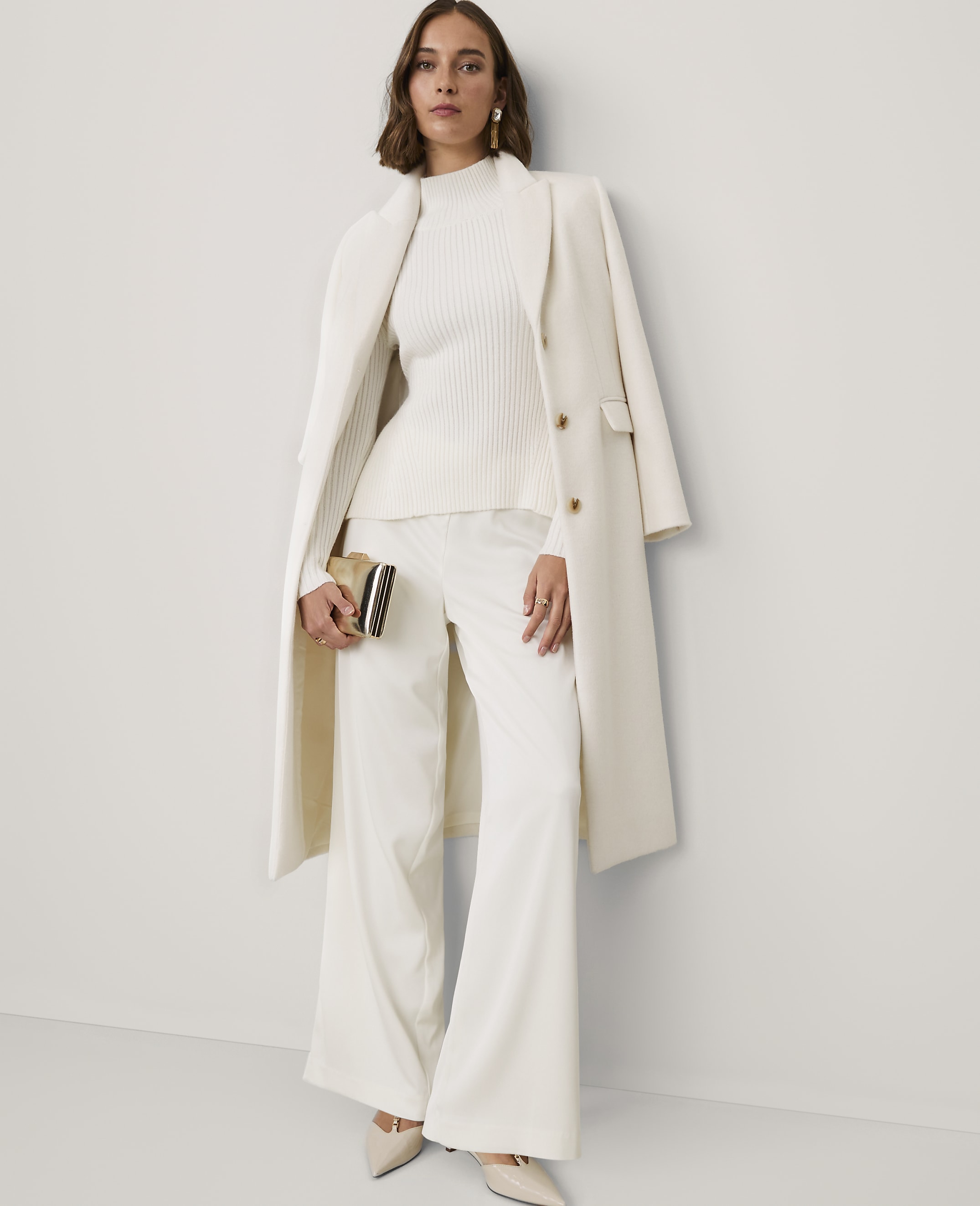 A woman wearing an all-white ensemble consisting of a ribbed sweater, wide-leg trousers, and a long coat stands against a plain white wall.