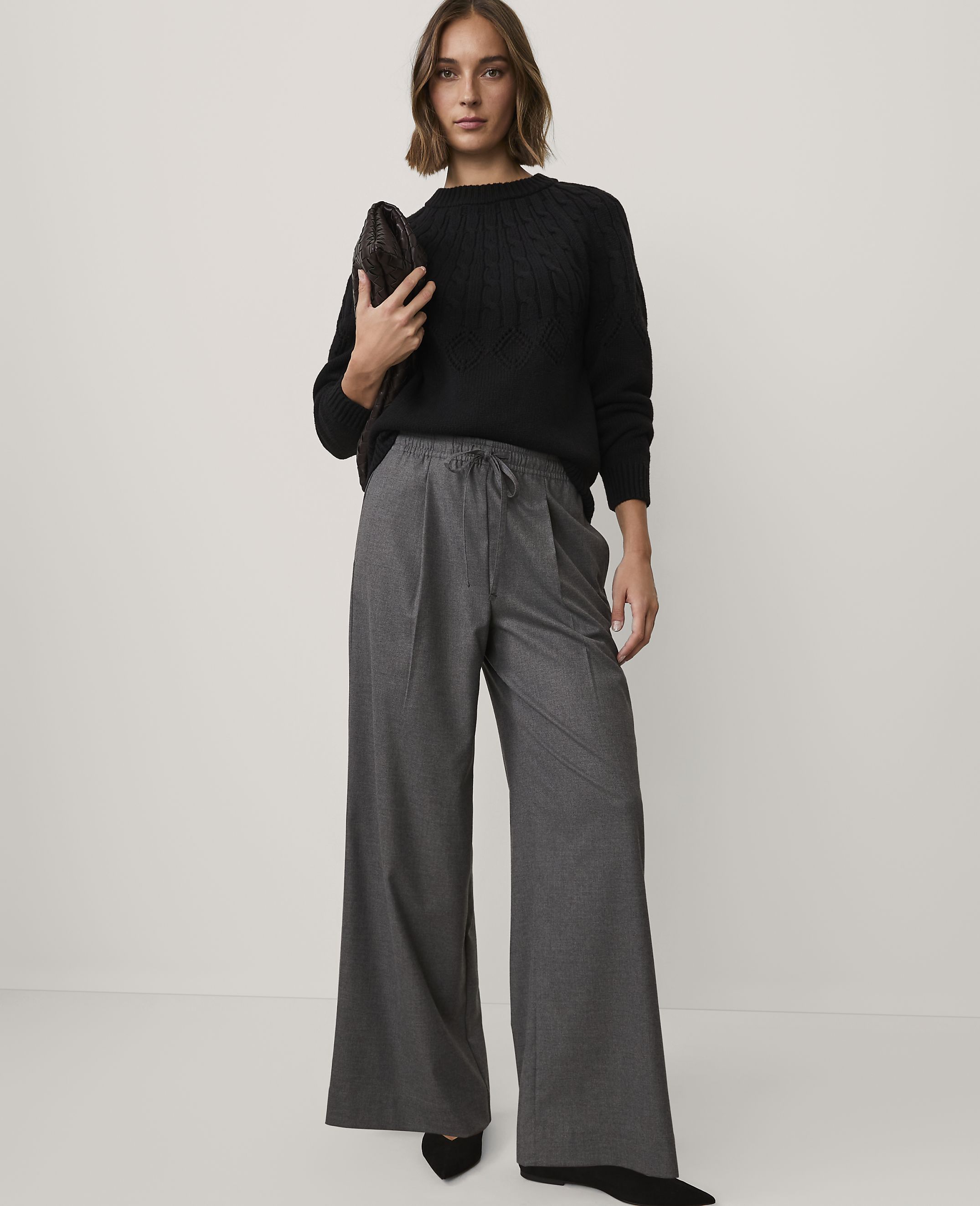 A woman wearing a black cable-knit sweater and wide-leg grey trousers stands against a plain white background.