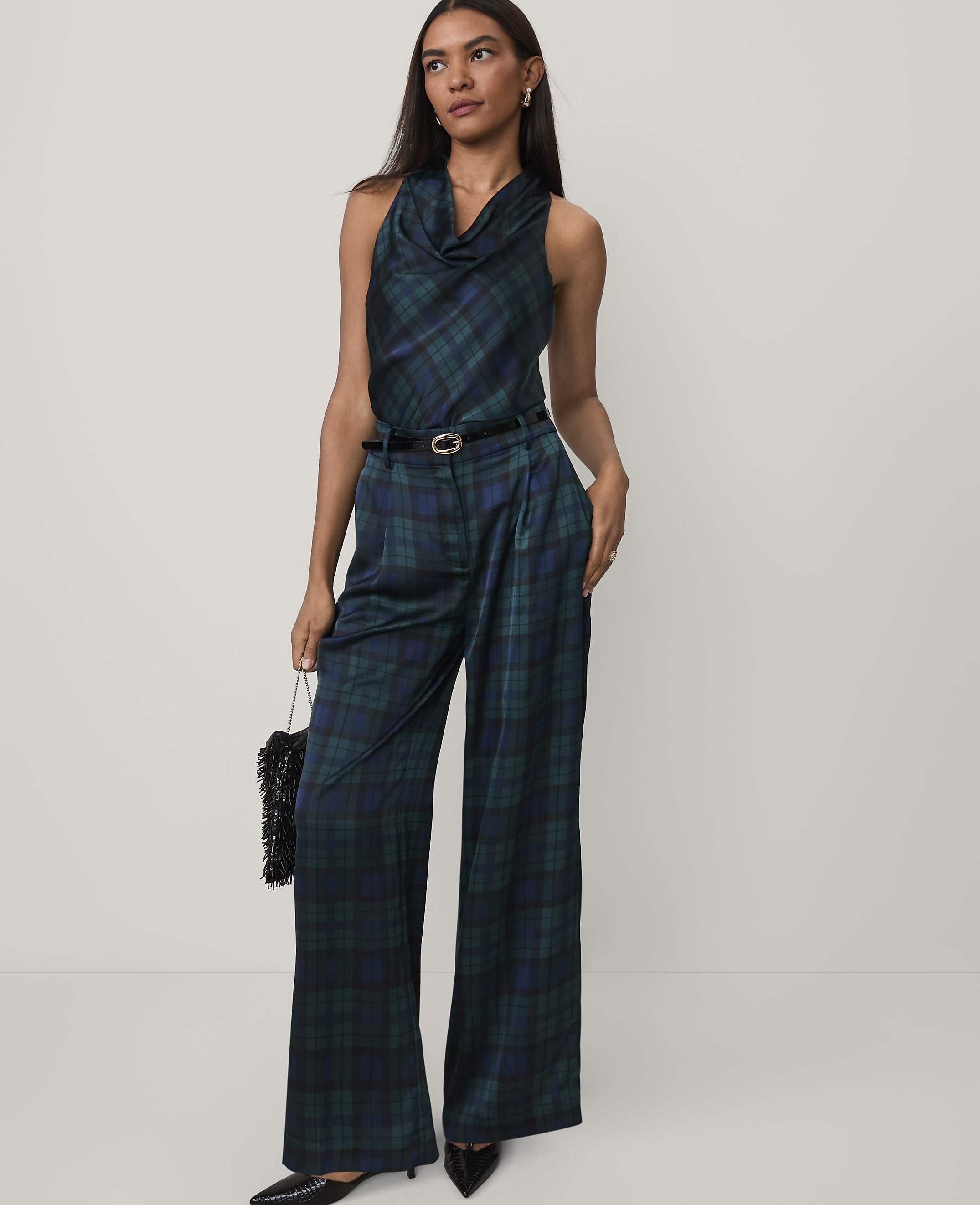 A woman wearing a dark green plaid jumpsuit and black heels stands against a plain white background.