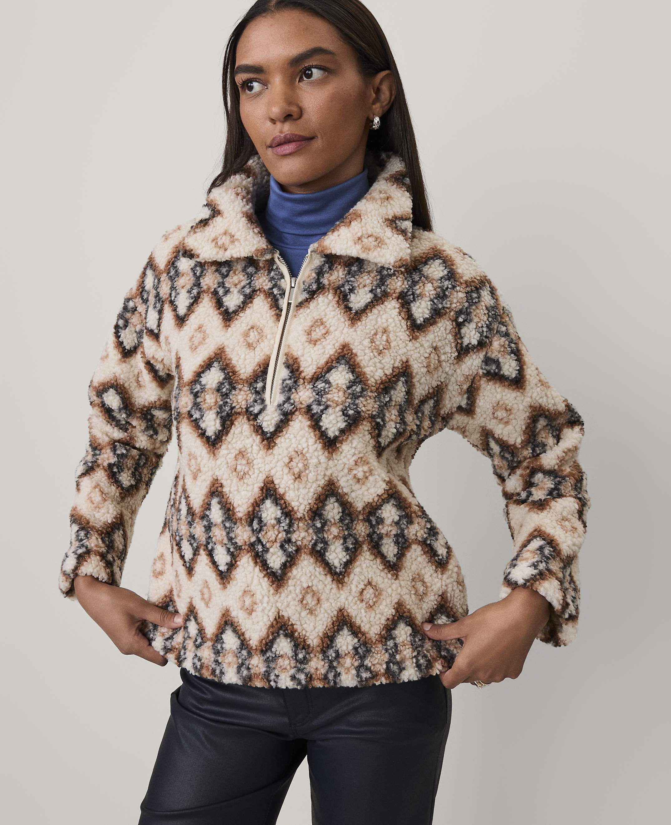 A woman wearing a patterned fleece pullover and black leather pants stands against a plain light gray background.