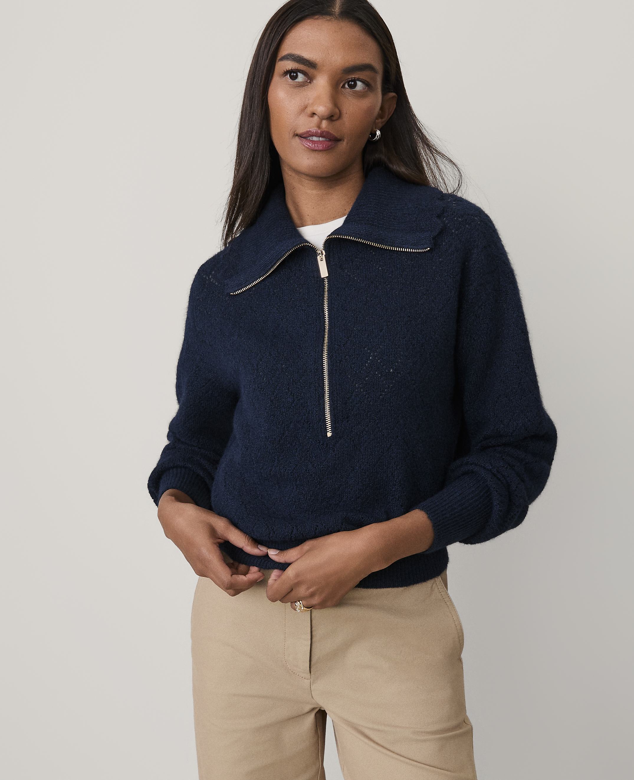 A woman wearing a navy blue quarter-zip sweater and khaki pants stands against a plain, light-colored background.