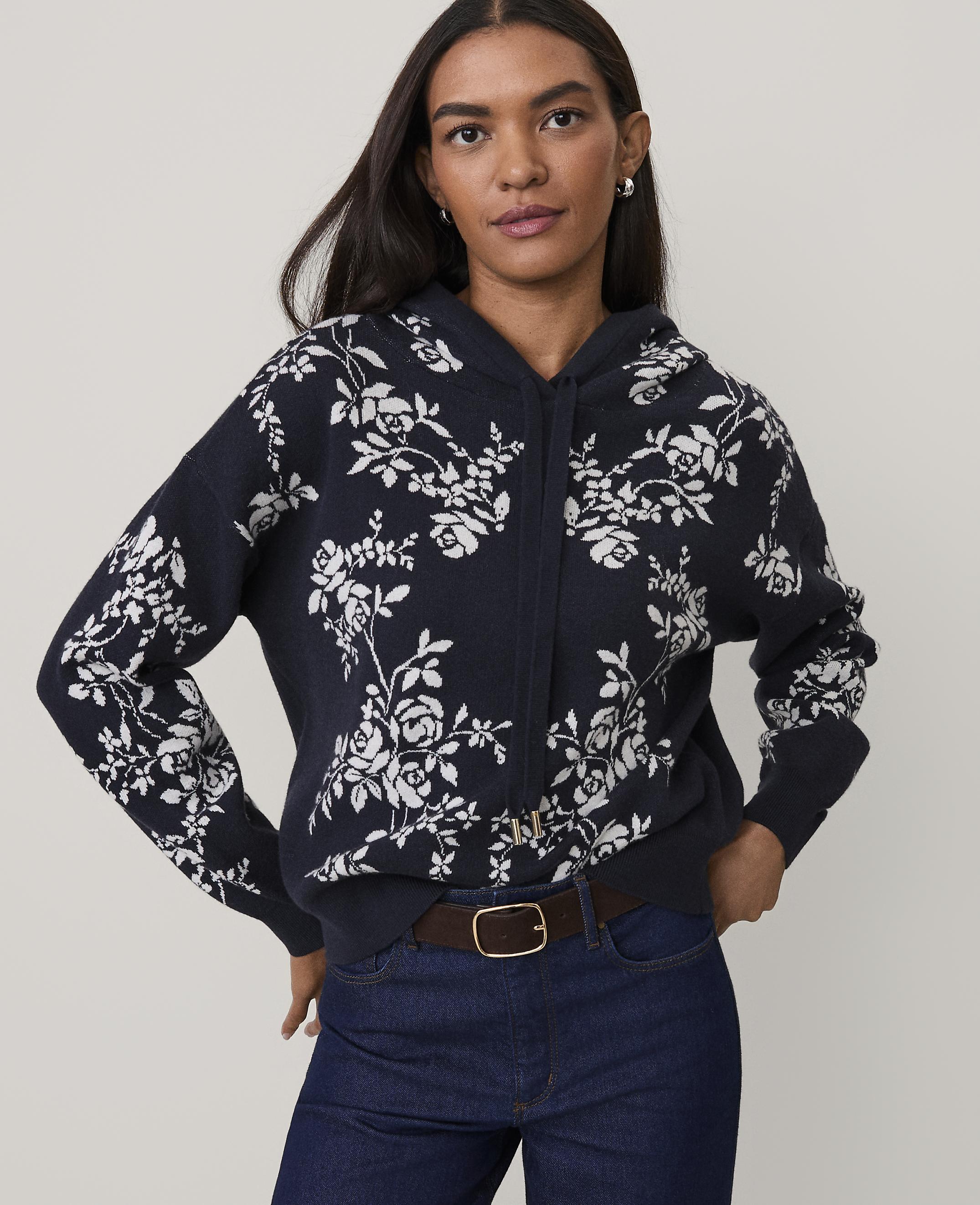 A woman wearing a navy blue floral hoodie and dark blue jeans stands against a plain, light-colored background.