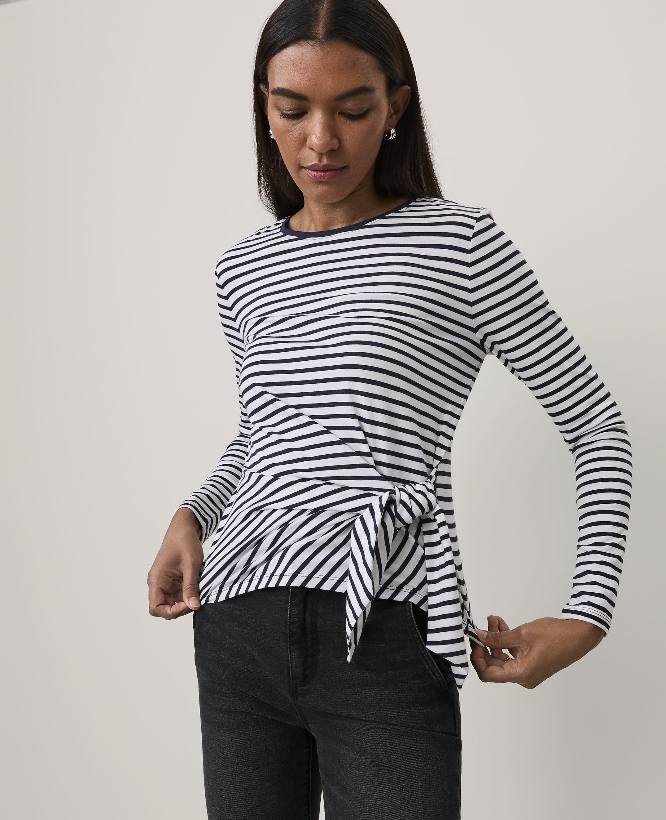 A woman wearing a navy and white striped long-sleeve top with a tie-front detail and black jeans stands against a plain white background.