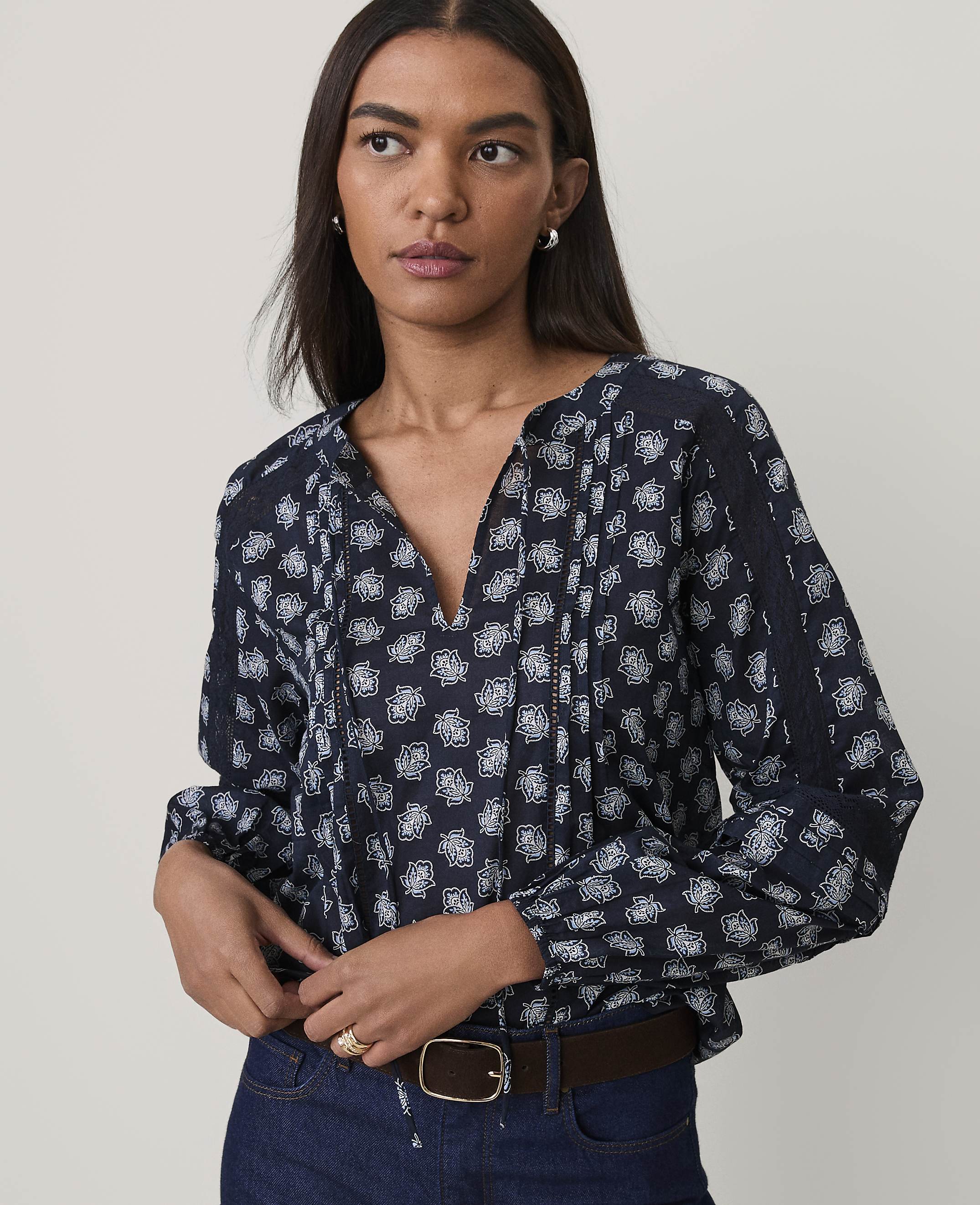A woman wearing a navy blue floral print blouse and blue jeans stands against a plain, light-colored background.