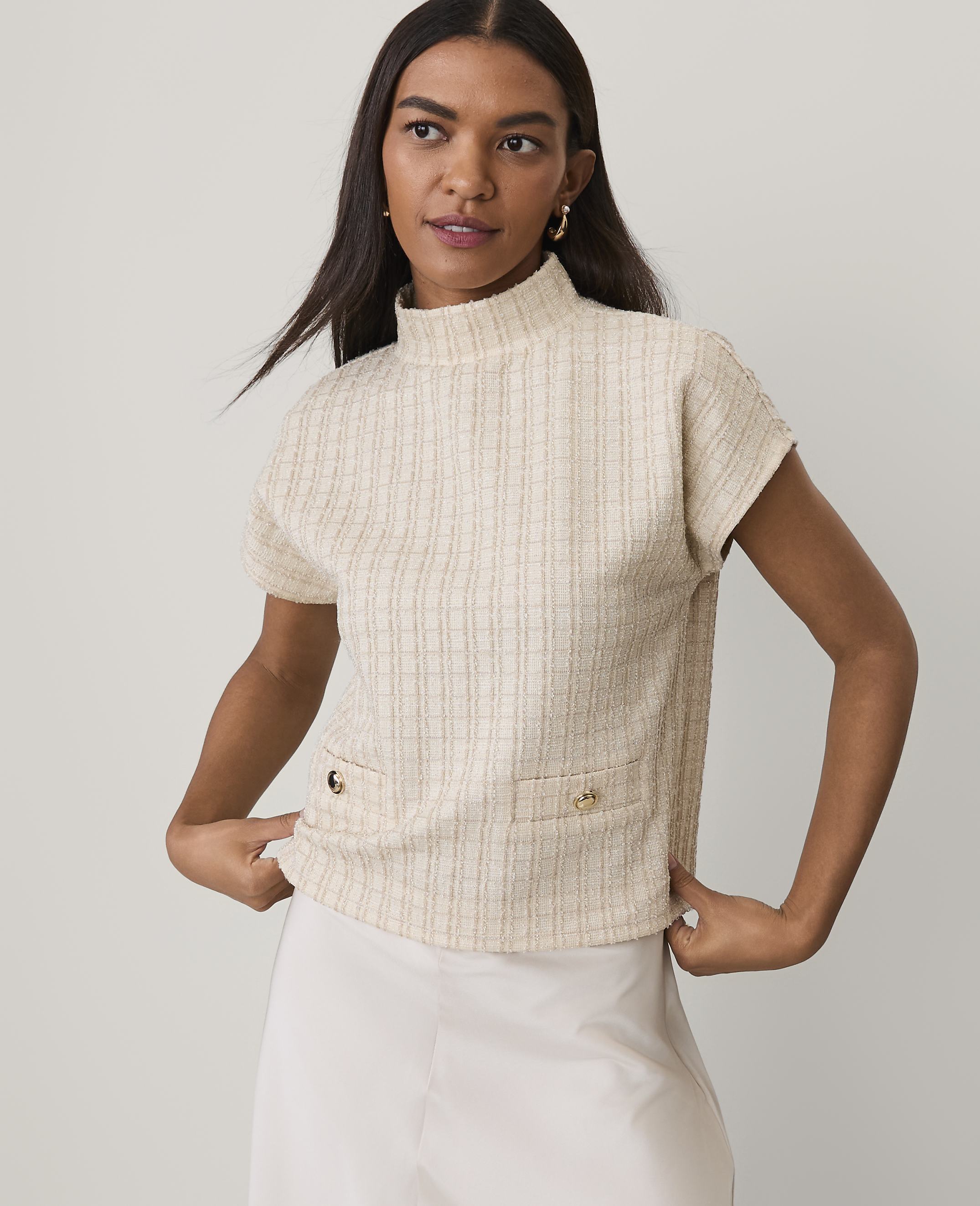 A woman wearing a cream-colored textured short-sleeve top and a white skirt stands against a plain light gray background.