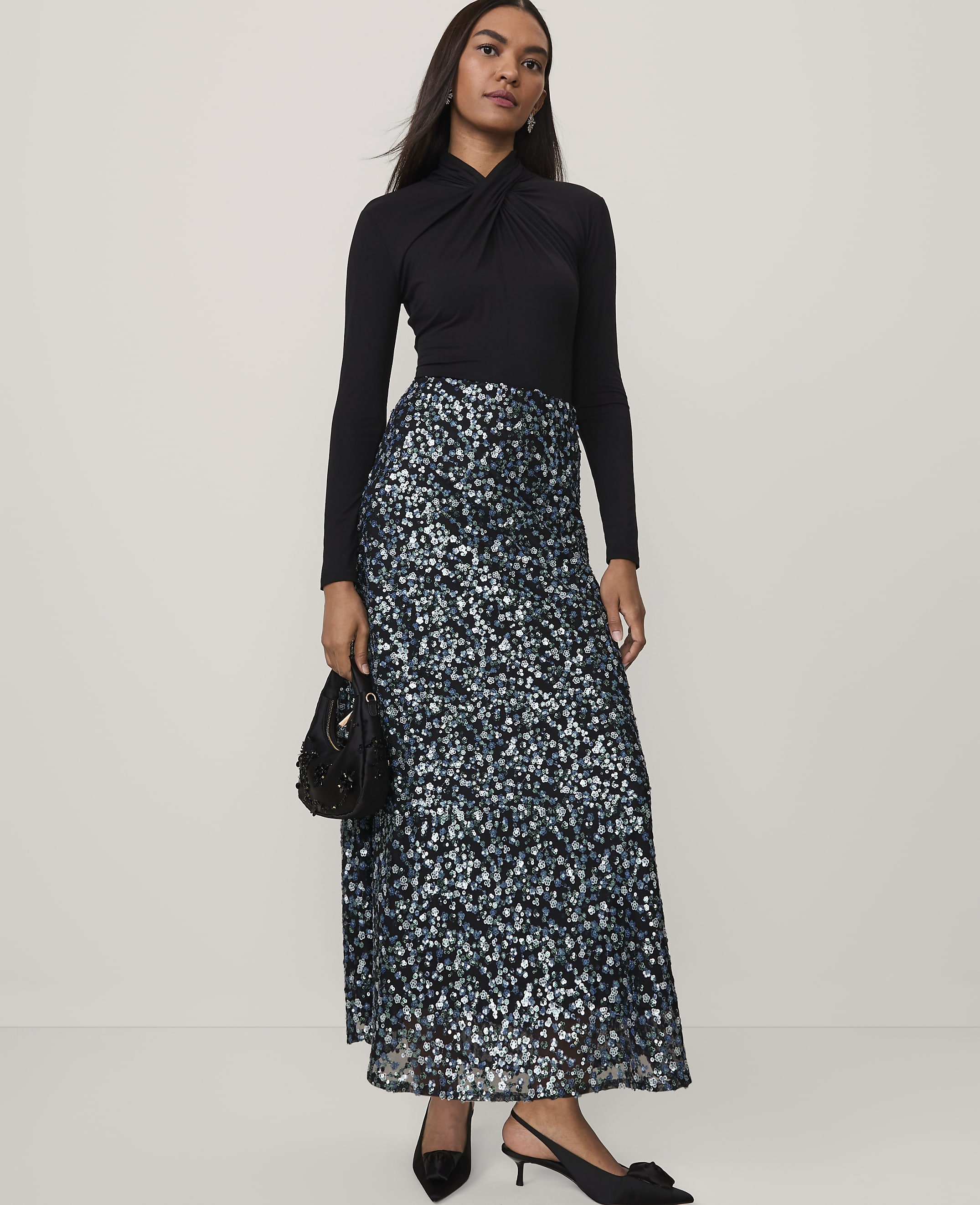 A woman wearing a black long-sleeved top and a shimmering blue and silver sequined skirt stands against a plain, light-colored background.