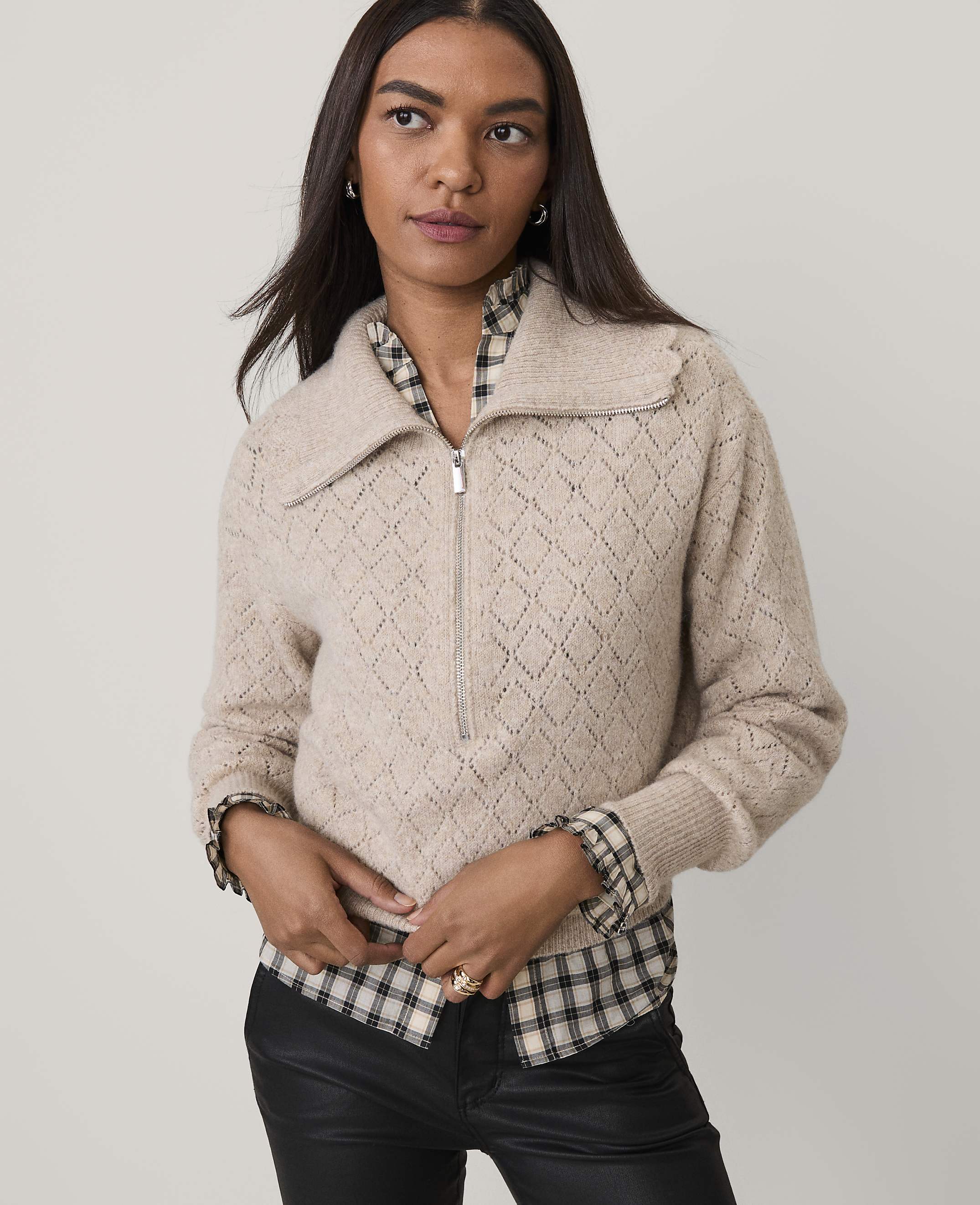 A woman wearing a beige textured zip-up sweater over a plaid shirt and black leather pants stands against a plain light gray background.
