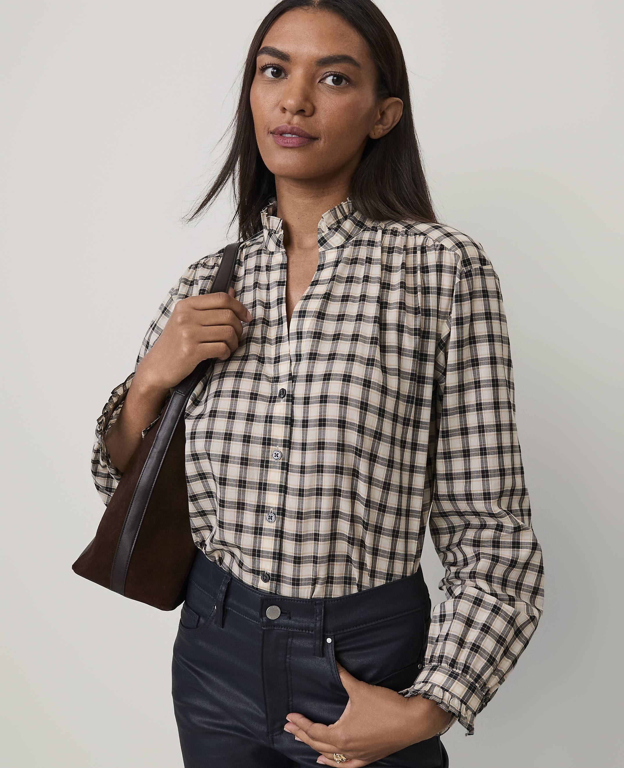 A woman wearing a plaid shirt and dark pants stands against a plain, light-colored background.