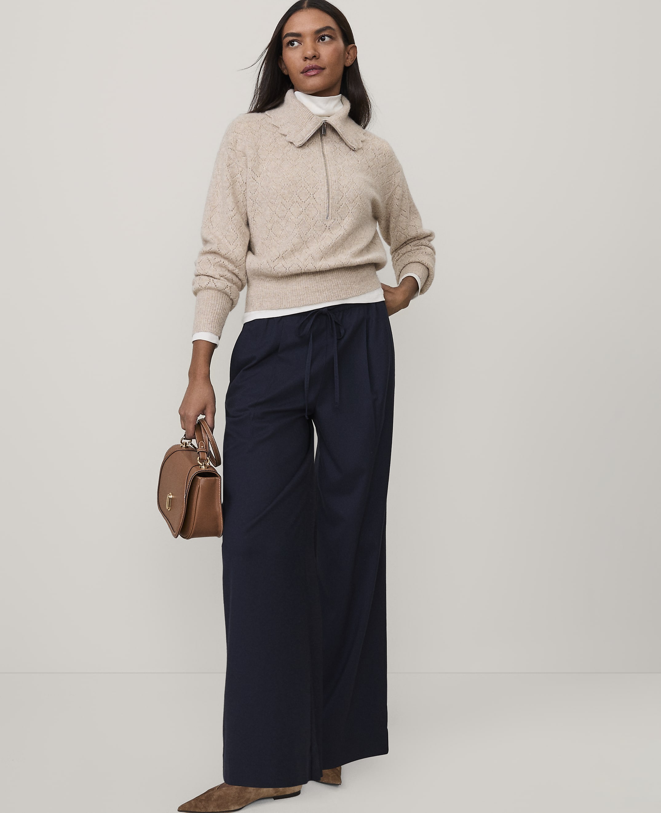 A woman wearing a beige sweater and navy wide-leg trousers stands against a plain white background.