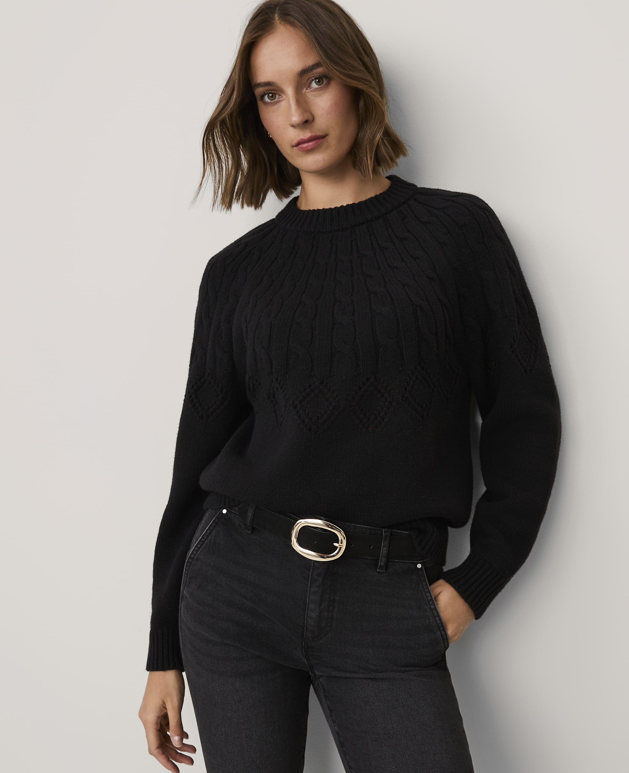 A woman wearing a black cable-knit sweater and black jeans with a gold buckle belt stands against a plain white wall.