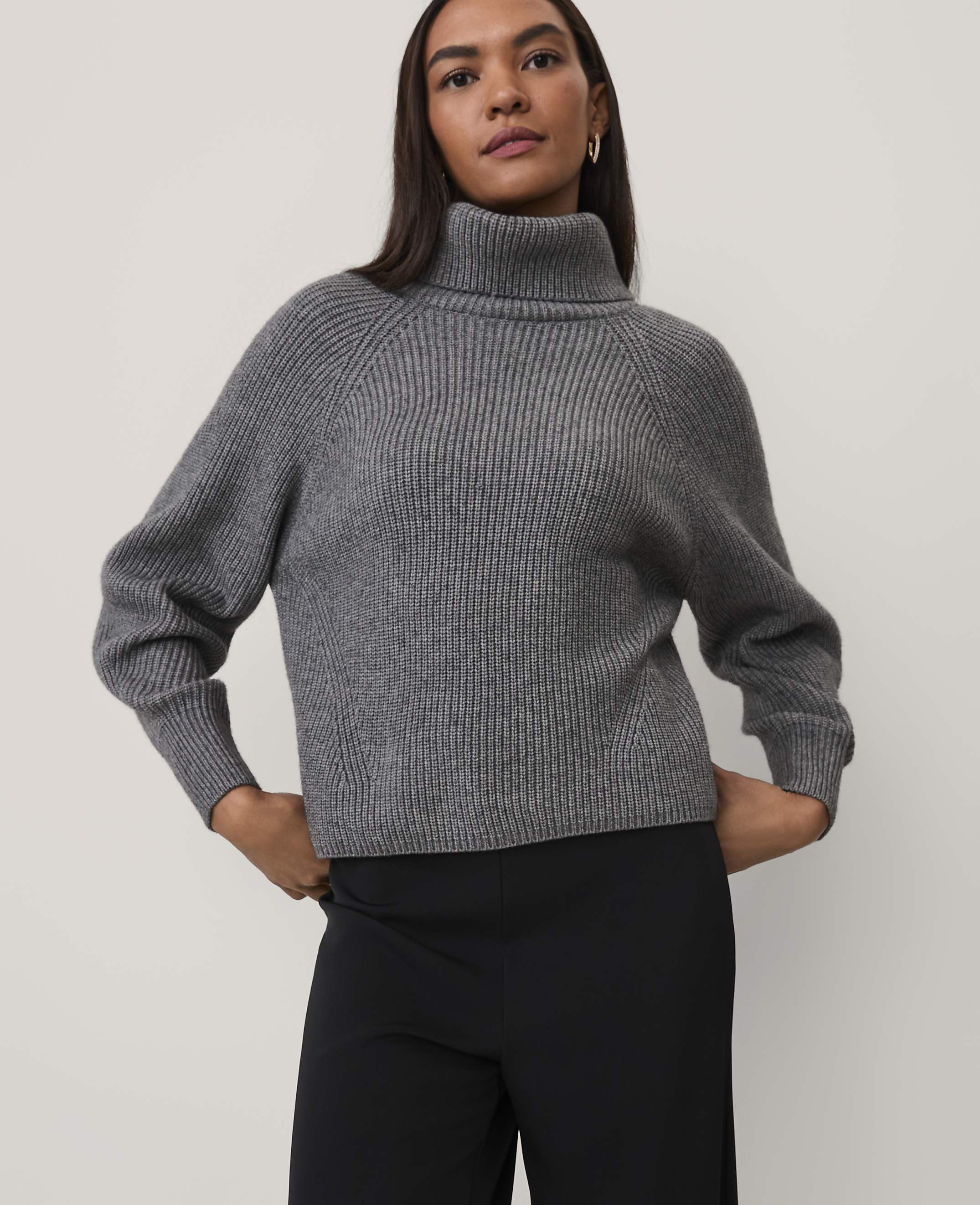 A woman wearing a grey ribbed turtleneck sweater and black trousers stands against a plain light grey background.