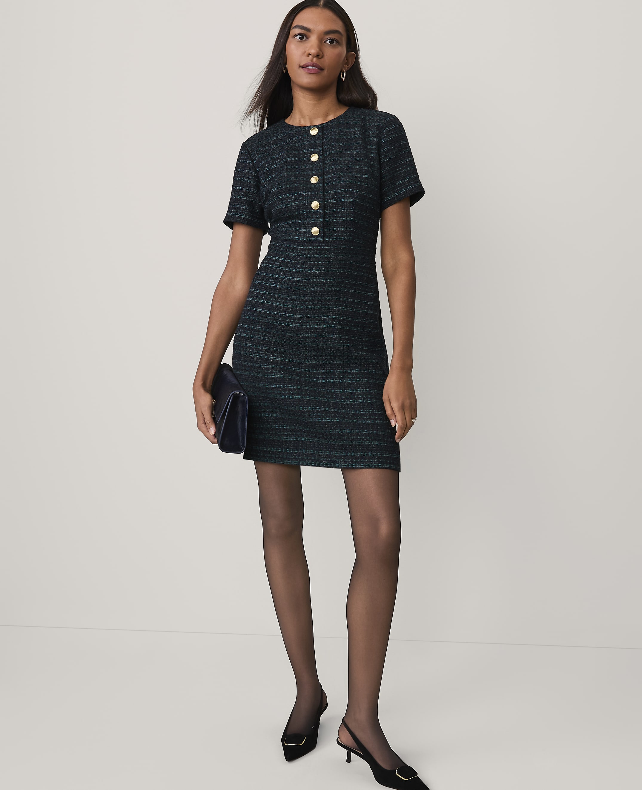 A woman wearing a dark green tweed dress and black heels stands against a plain white background.