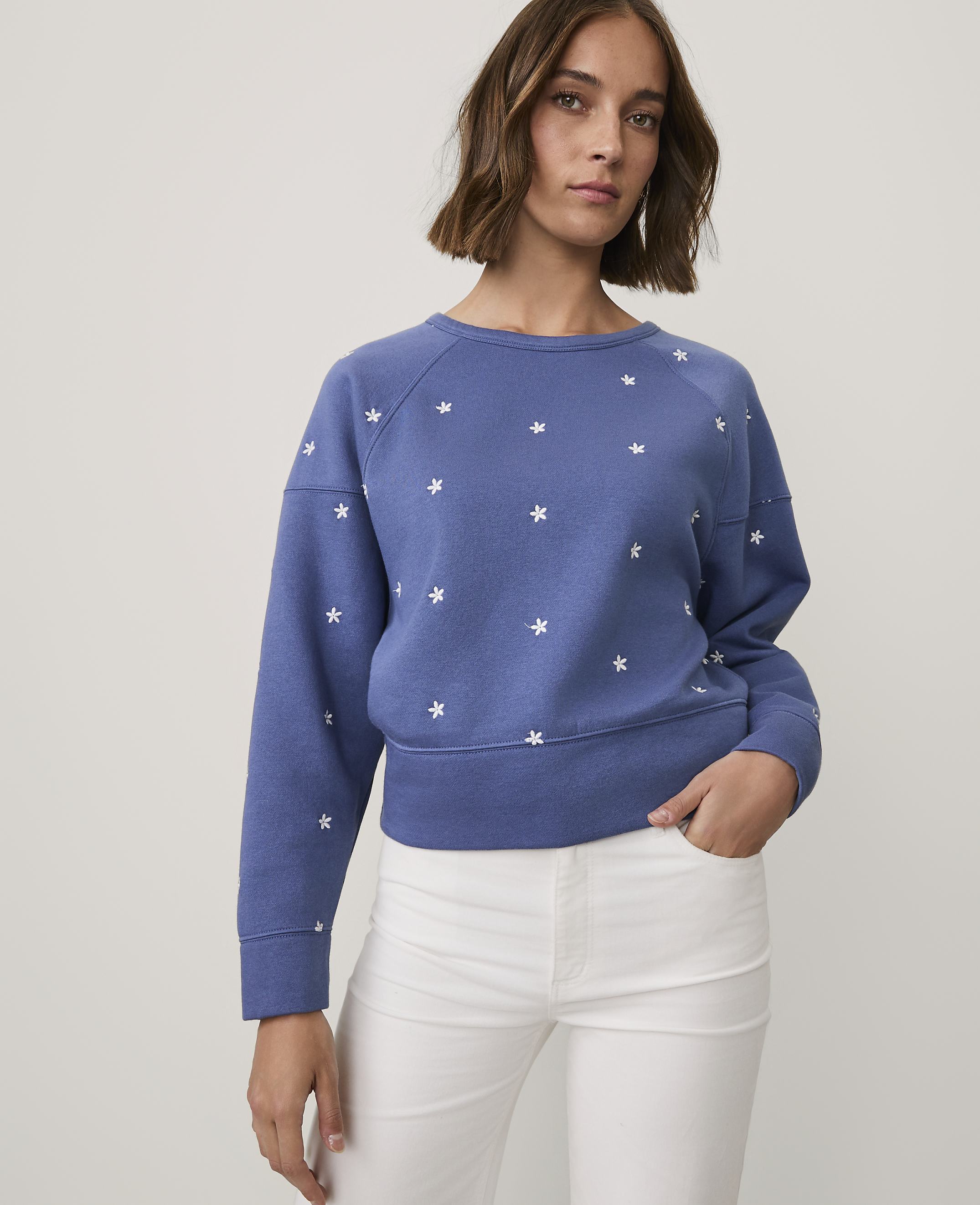 A woman wearing a blue star-patterned sweatshirt and white pants stands against a plain light gray background.