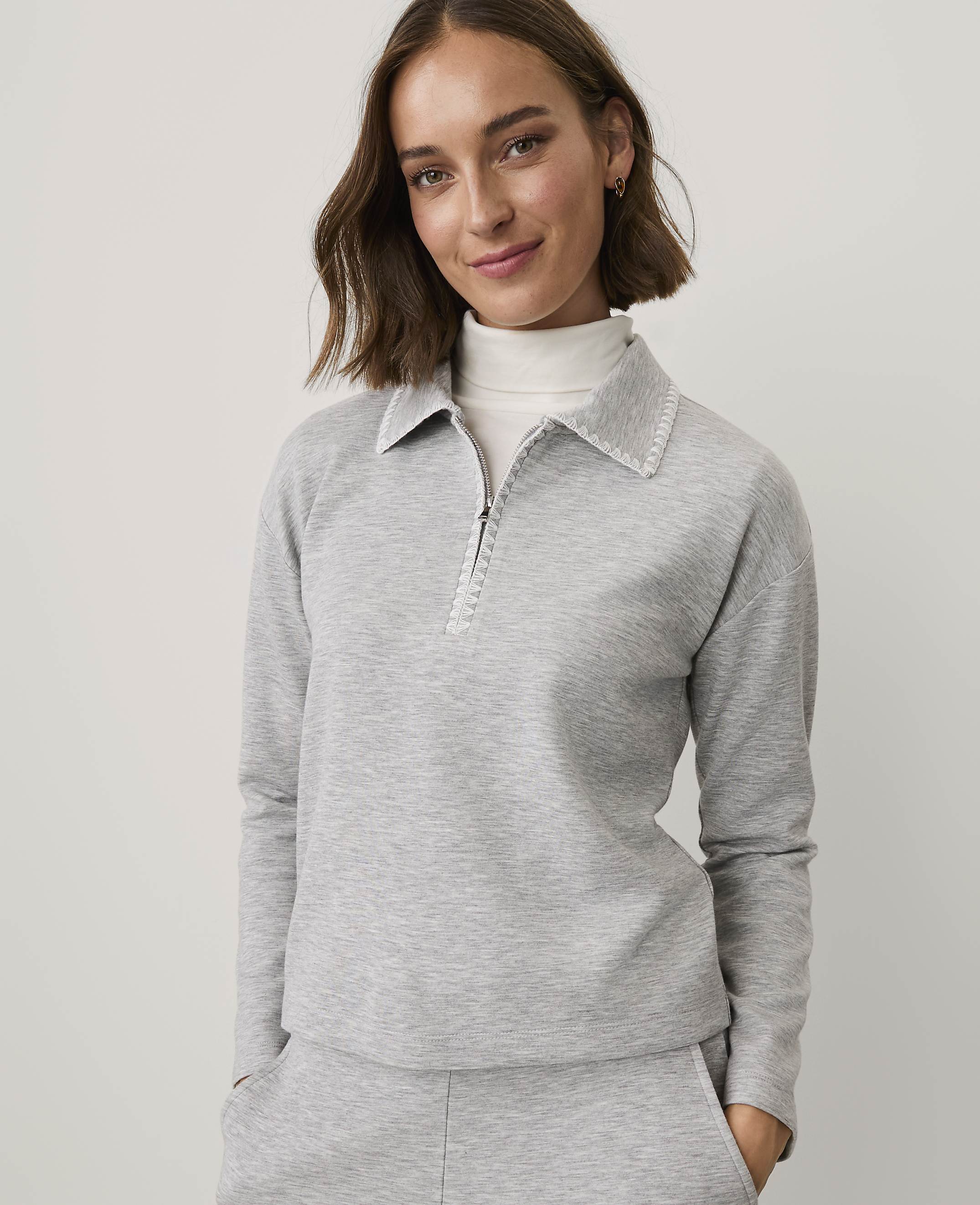A woman wearing a grey quarter-zip pullover and matching pants stands against a plain, light-colored background.