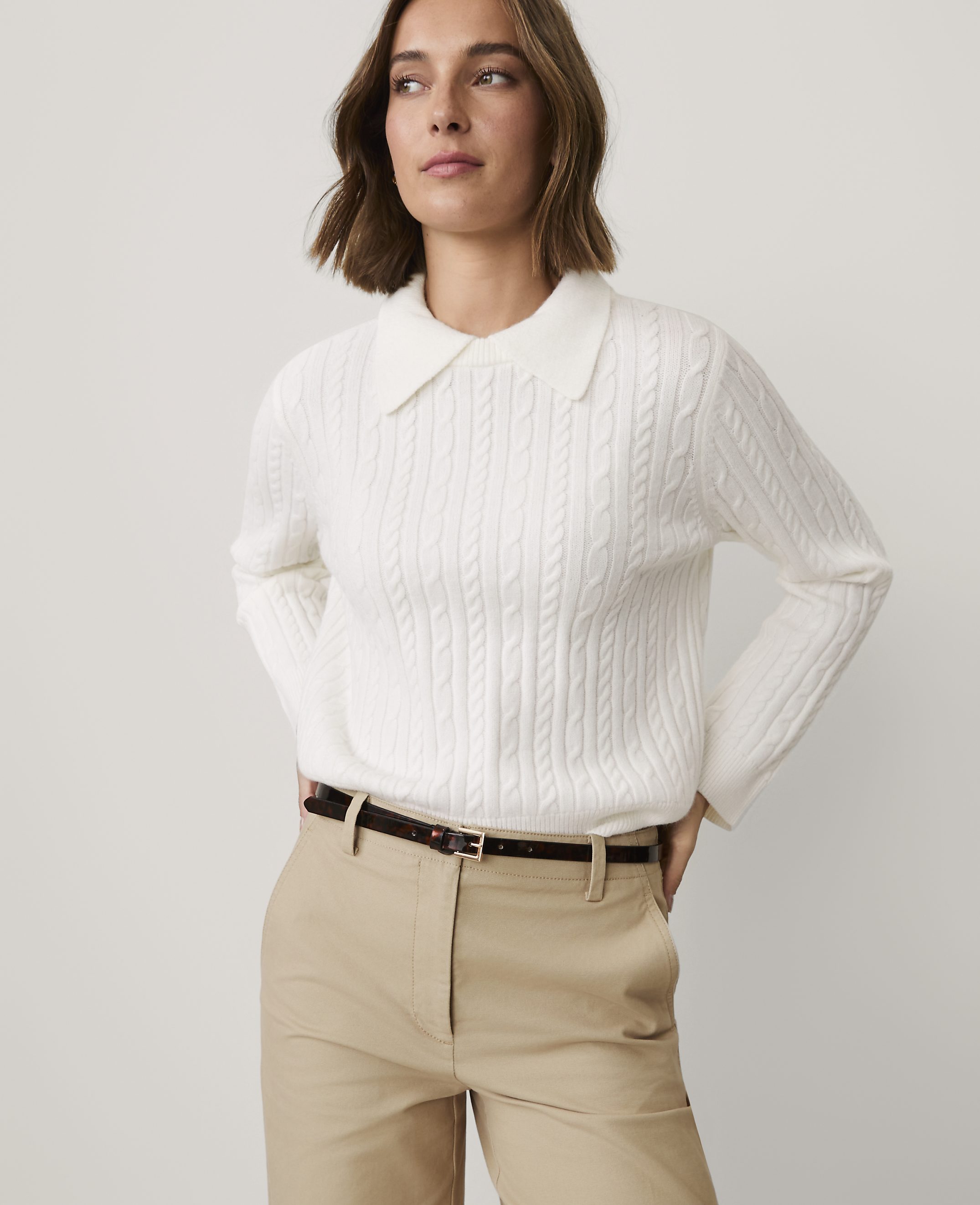 A woman wearing a white cable-knit collared sweater and khaki pants stands against a plain light gray background.