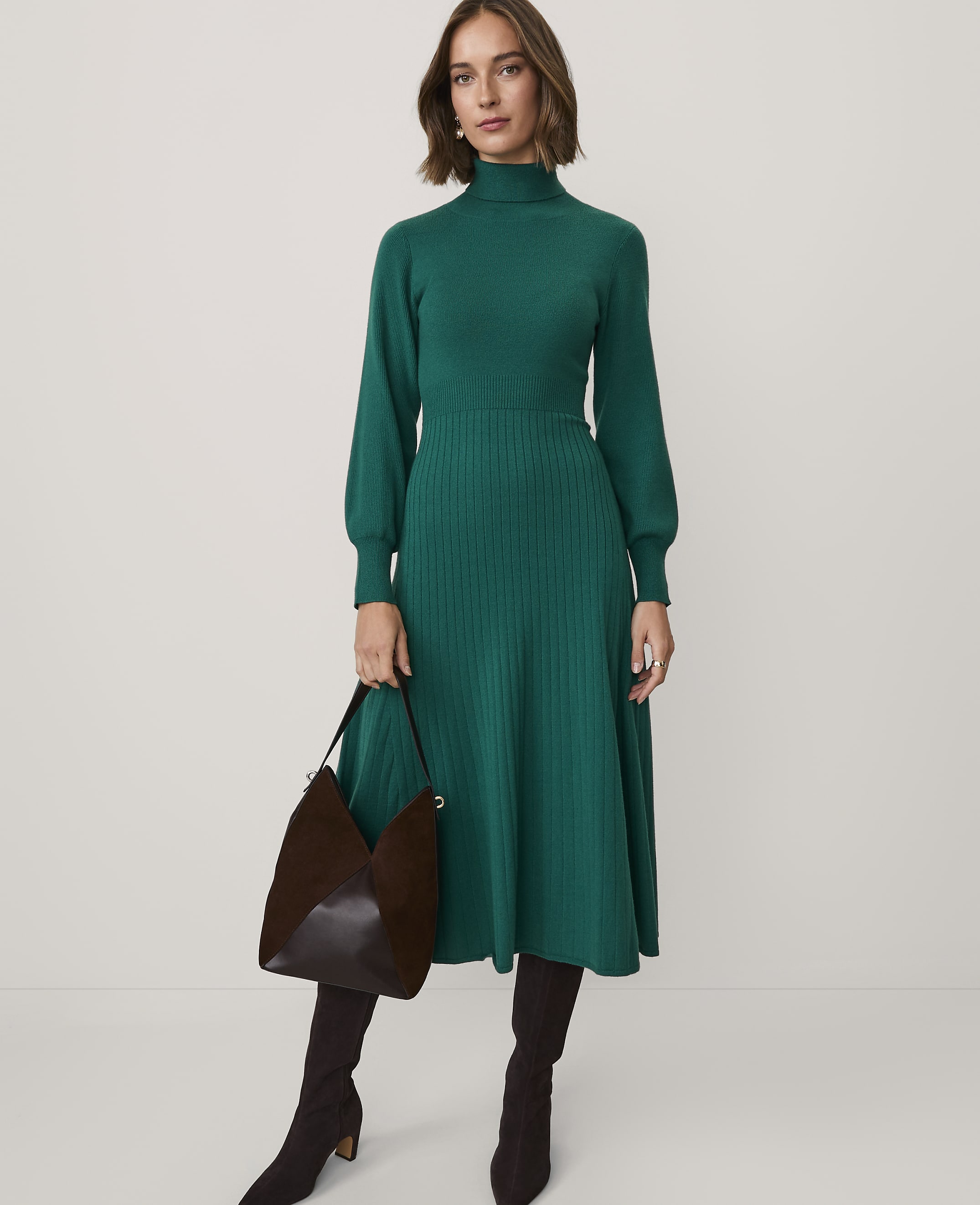 A woman wearing a long green knit dress and brown boots stands against a plain white background.