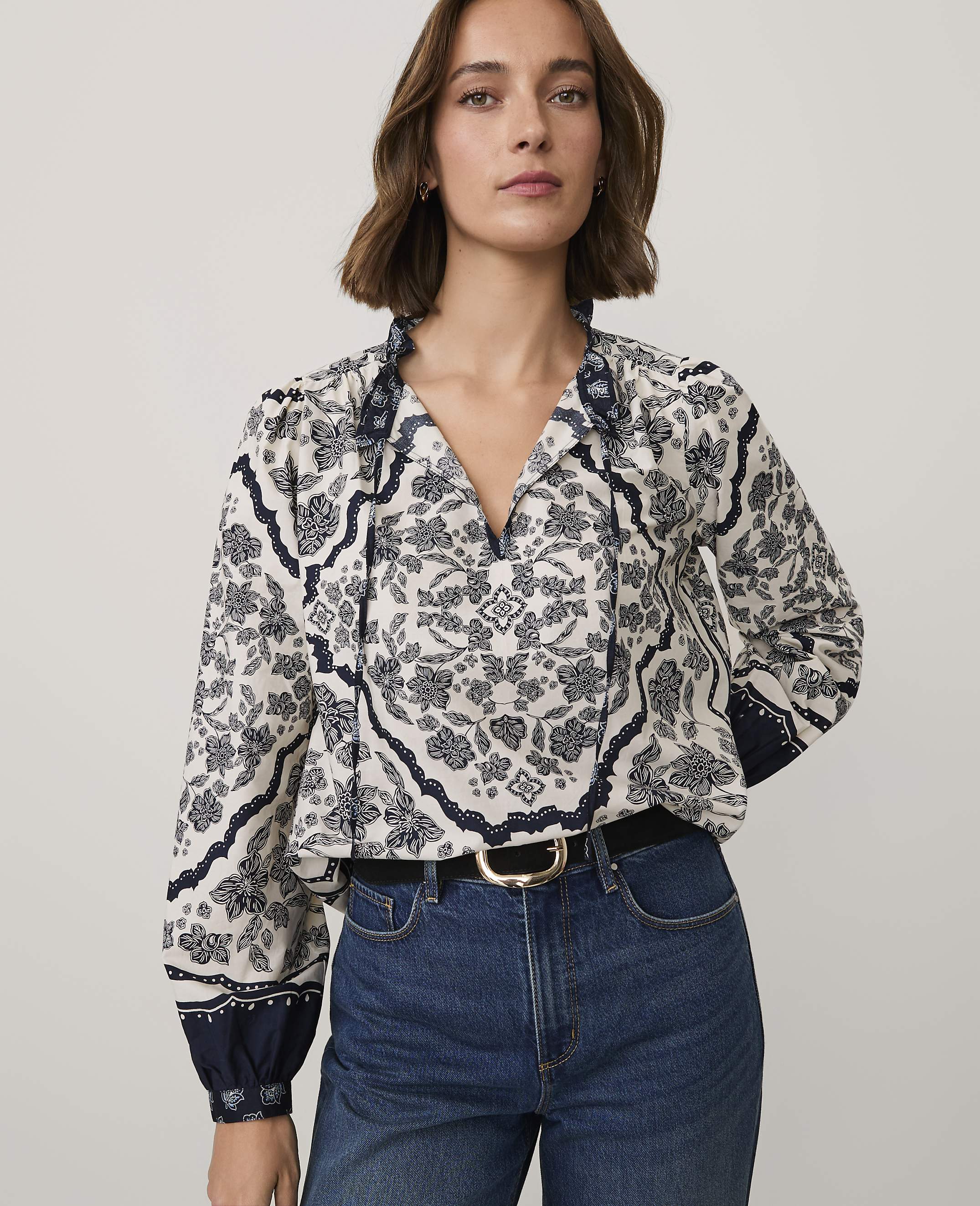 A woman wearing a patterned white and navy blouse and blue jeans stands against a plain light gray background.
