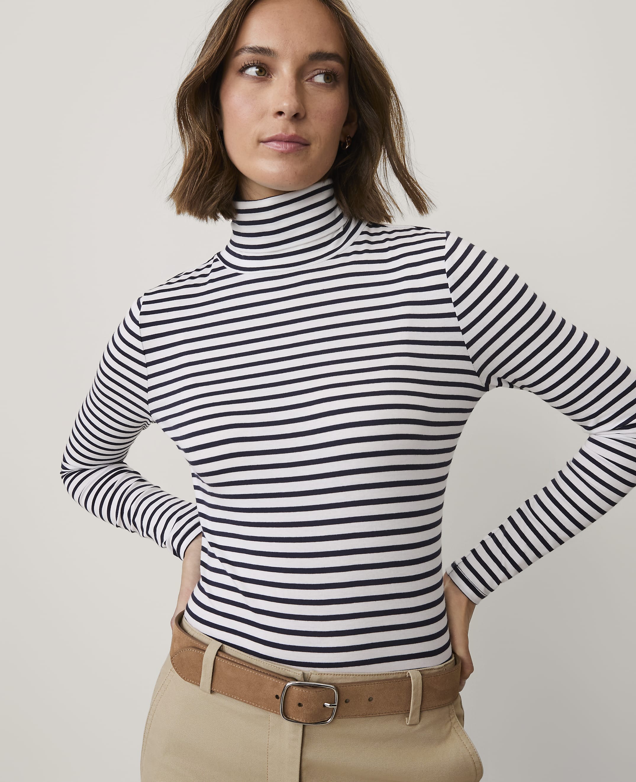 A woman wearing a navy and white striped turtleneck and khaki pants stands against a plain light gray background.
