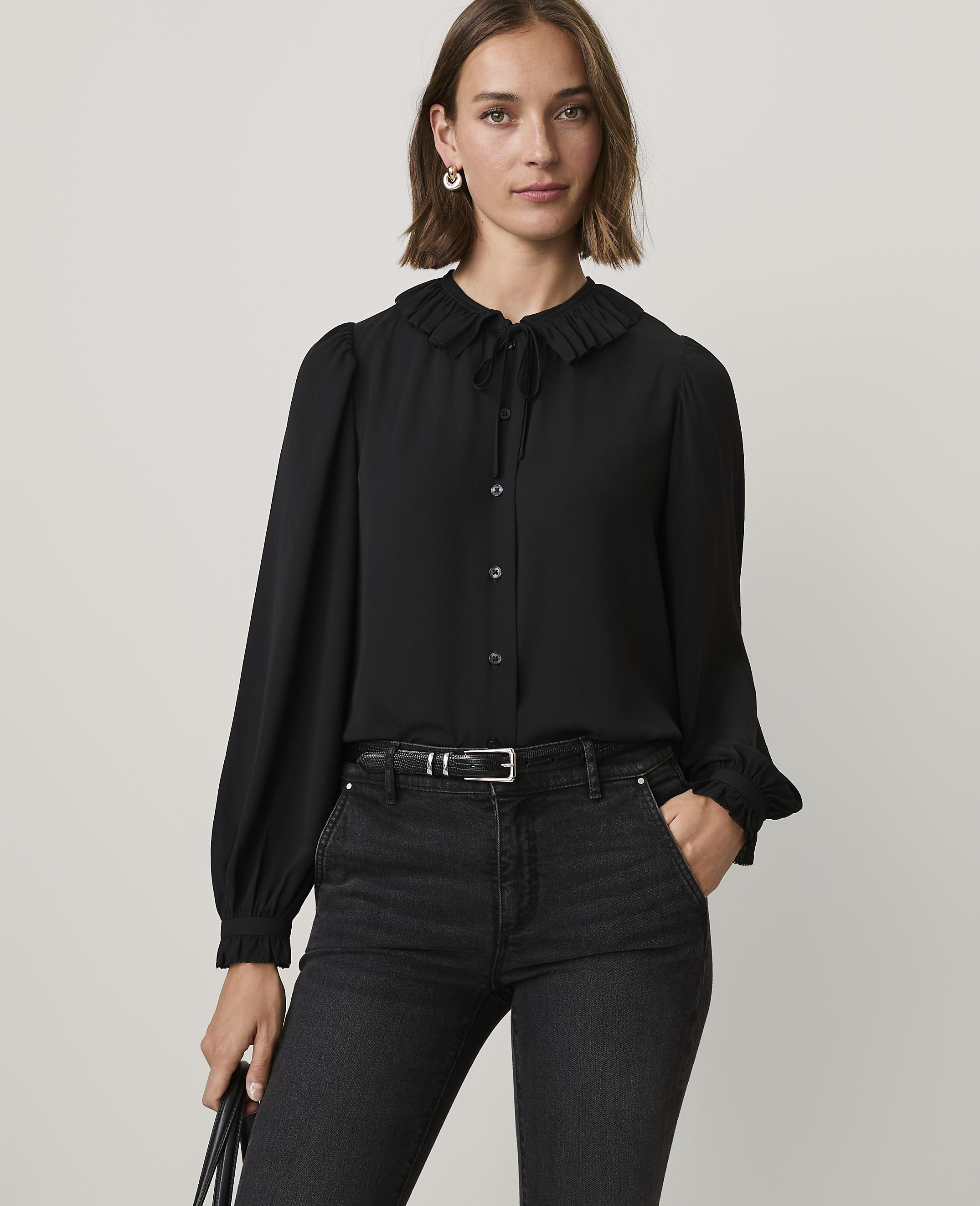 A woman wearing a black ruffled blouse and black jeans stands against a plain, light-colored background.
