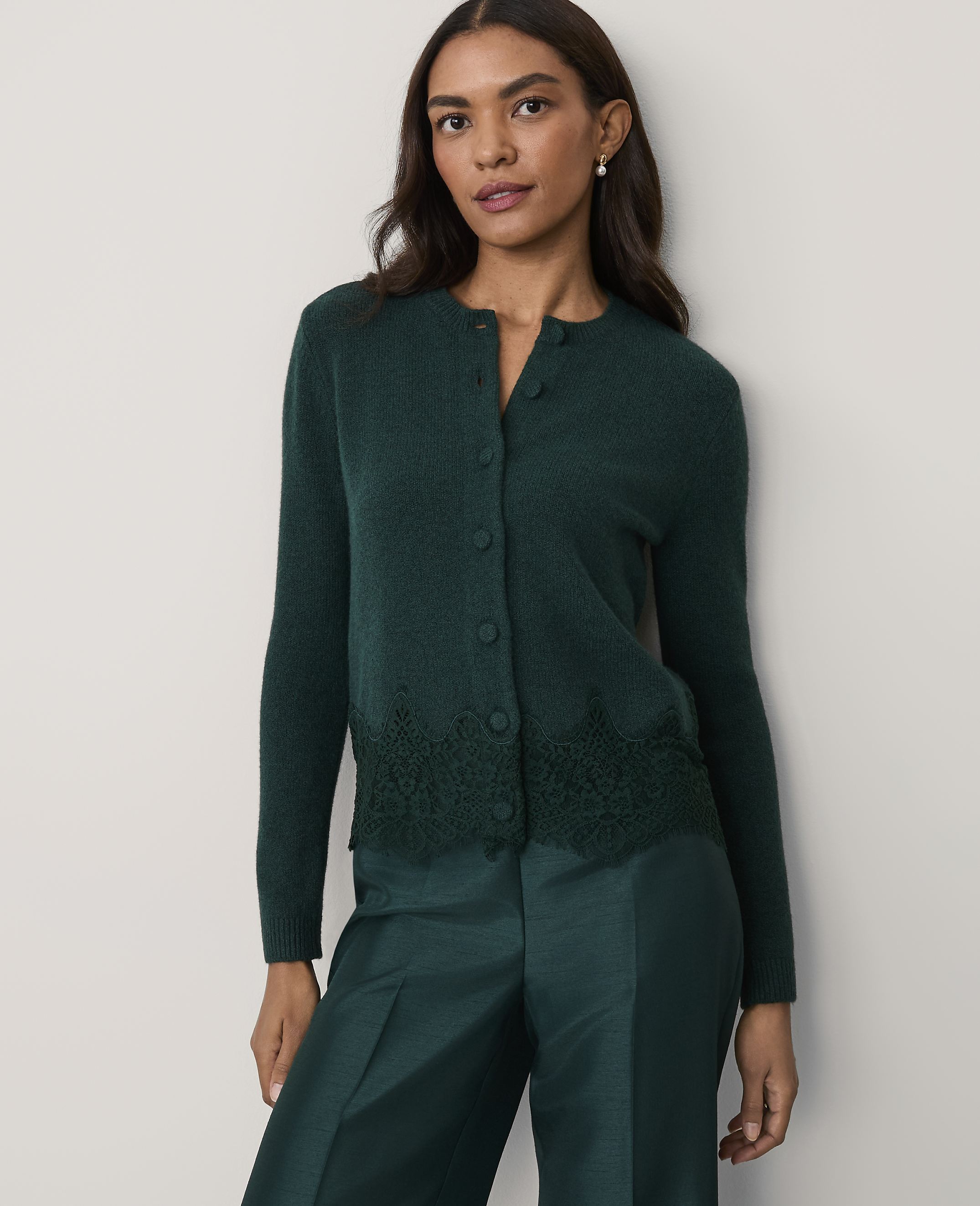 A woman wearing a dark green cardigan with lace detailing and matching trousers stands against a plain, light-colored wall.