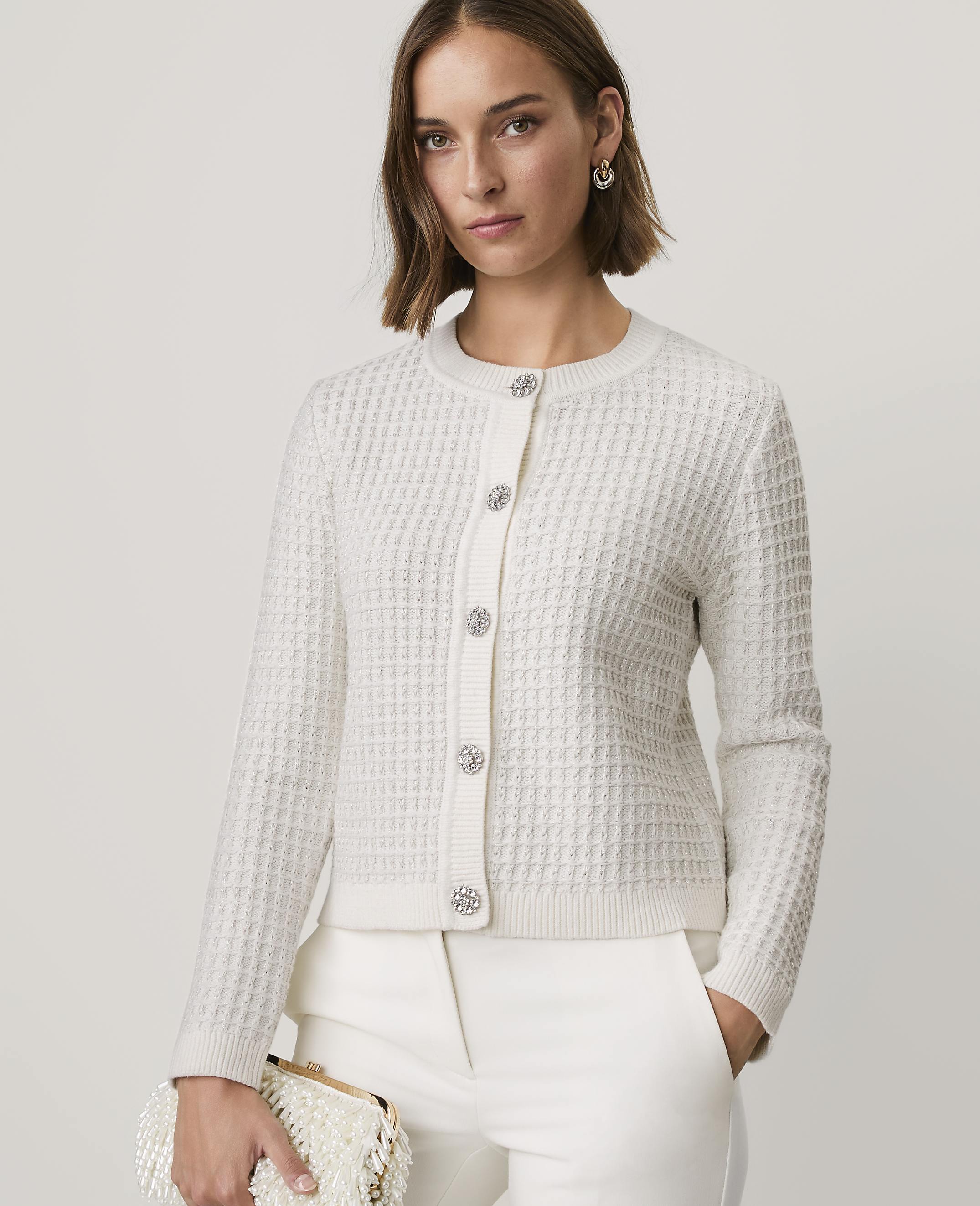 A woman wearing a white textured cardigan and white trousers stands against a plain, light-colored background.