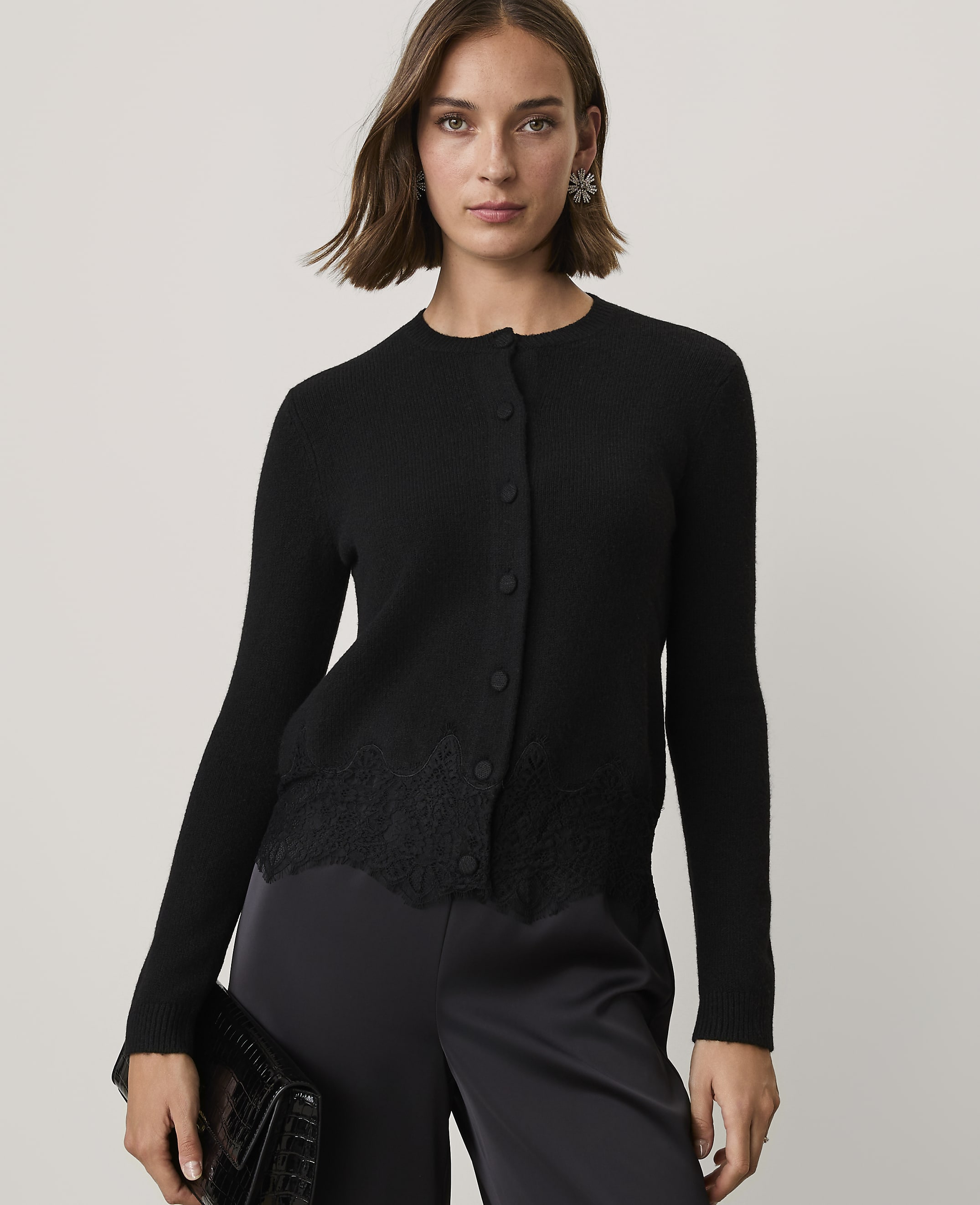 A woman wearing a black cardigan with lace detailing and black satin trousers stands against a plain light gray background.