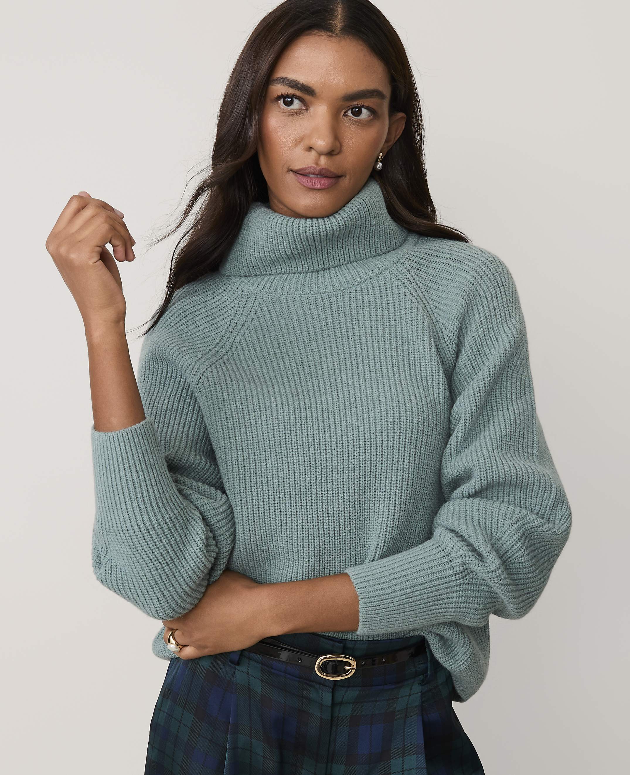 A woman wearing a light blue turtleneck sweater and a plaid skirt stands against a plain, light-colored background.