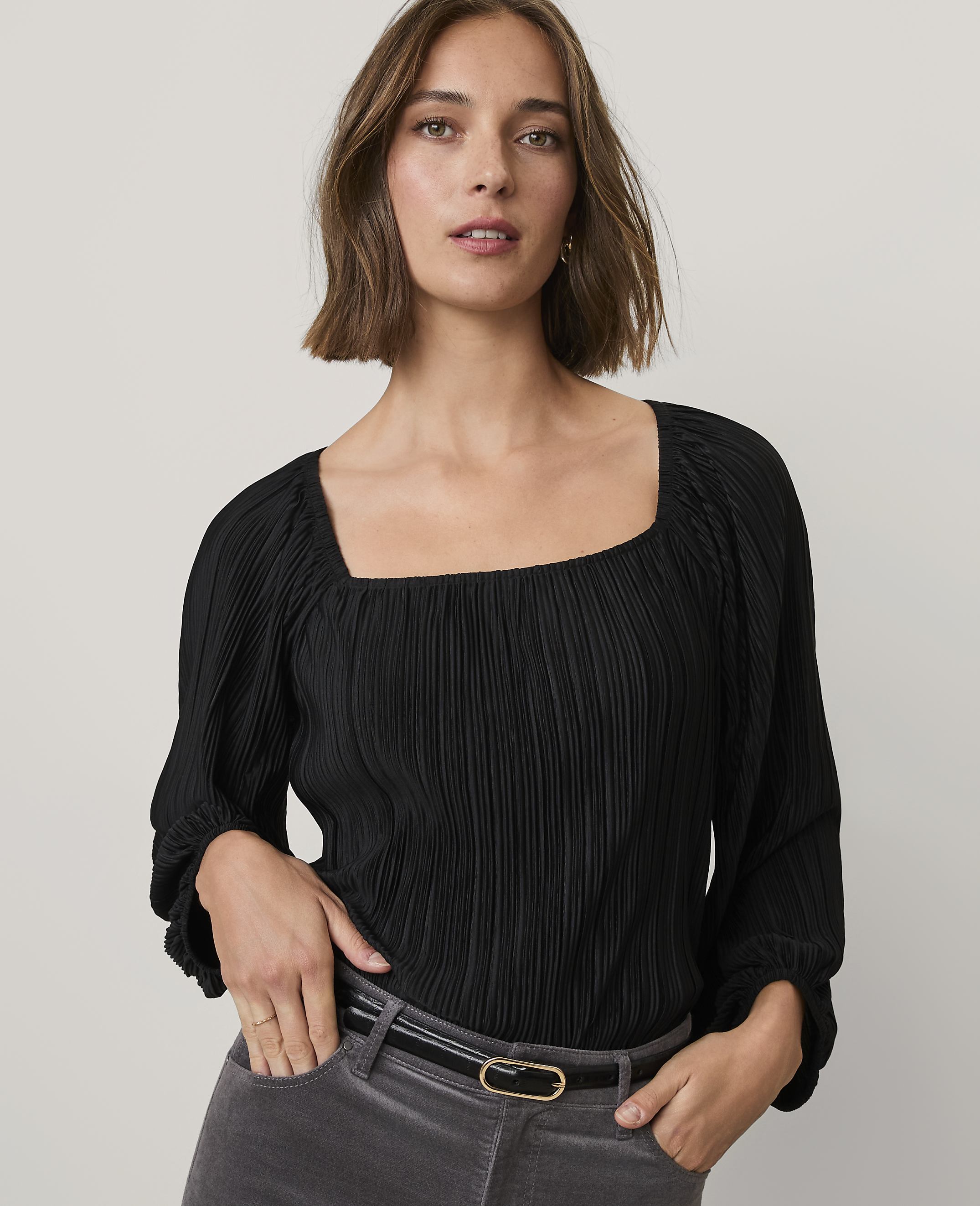 A woman wearing a black pleated top and grey trousers stands against a plain light grey background.