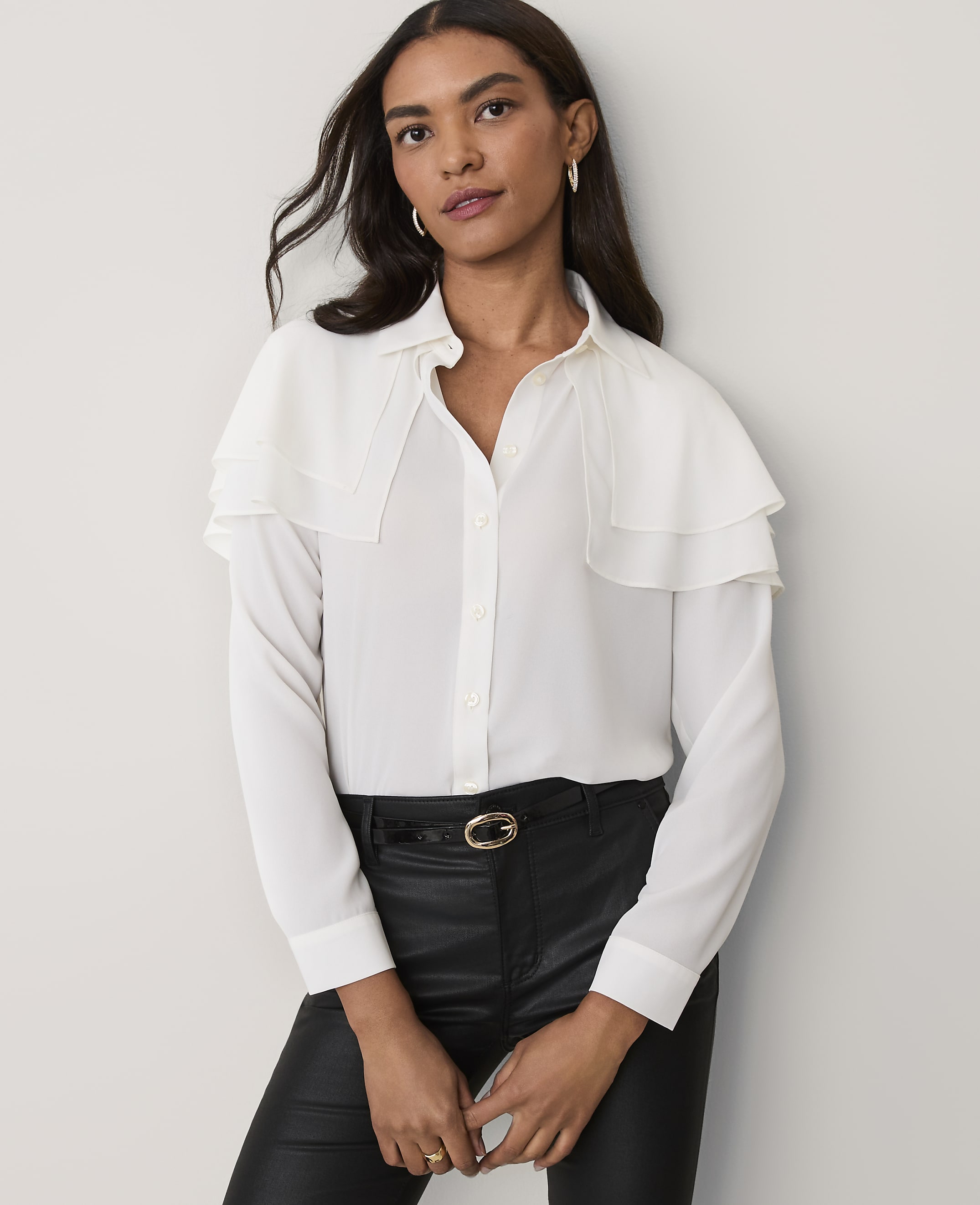 A woman wearing a white ruffled blouse and black leather pants stands against a plain light-grey background.