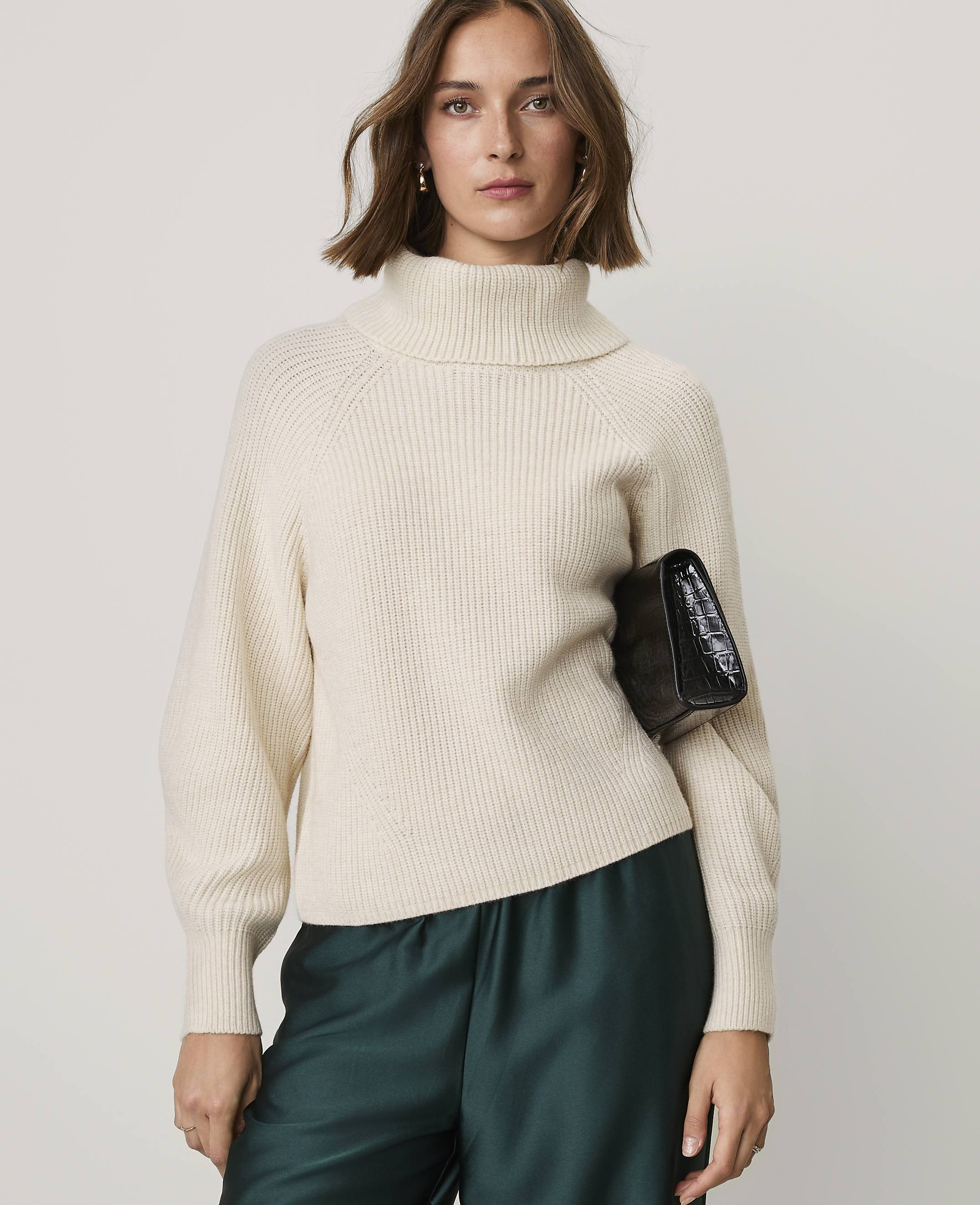 A woman wearing a cream-colored turtleneck sweater and dark green satin pants holds a black clutch against a plain light gray background.